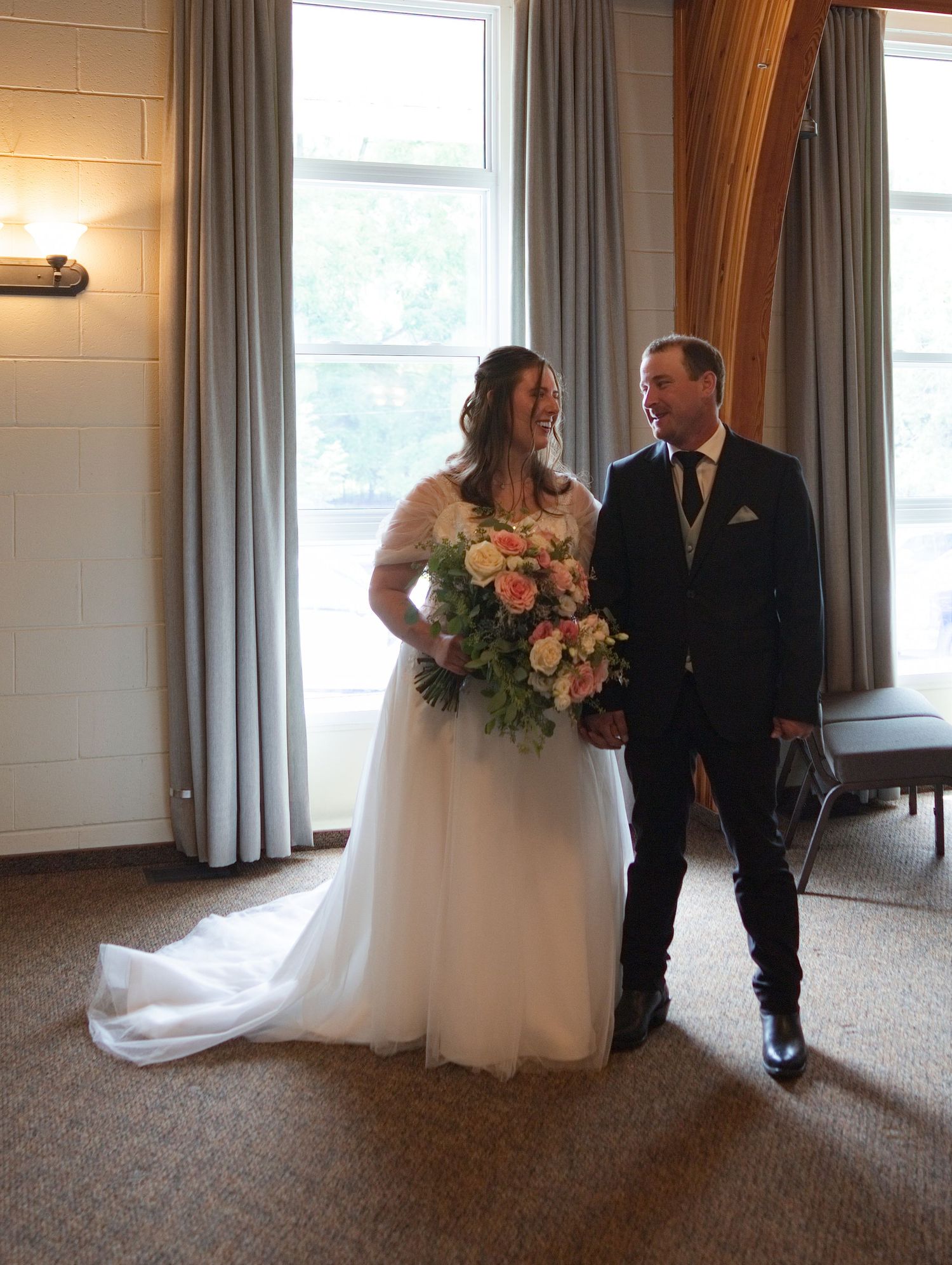 Bride in white gown holding pink bouquet stands with groom in dark suit near large windows at wedding venue.