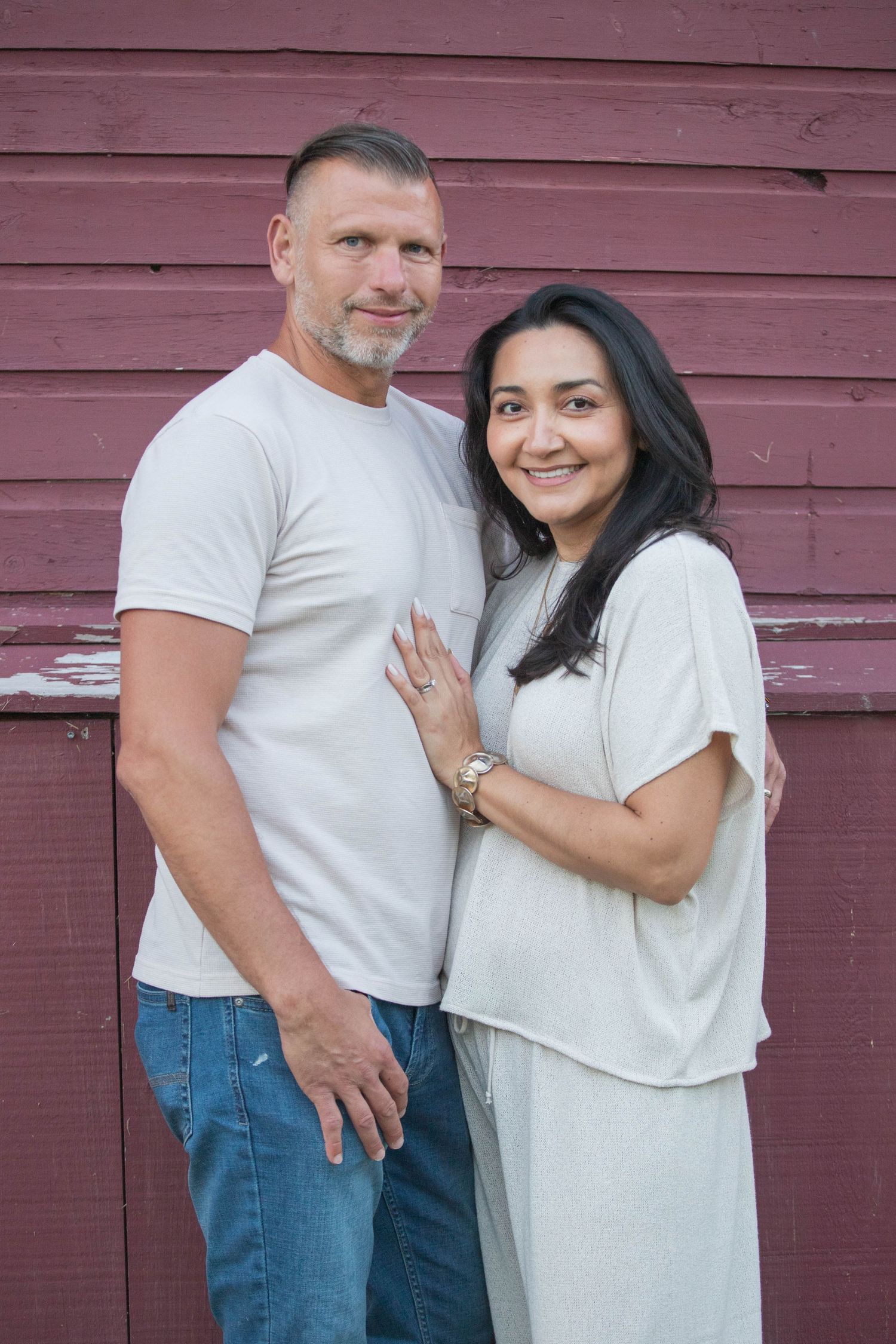 A middle-aged couple poses warmly against a rustic red barn wall, man in white tee and jeans, woman in light gray dress.