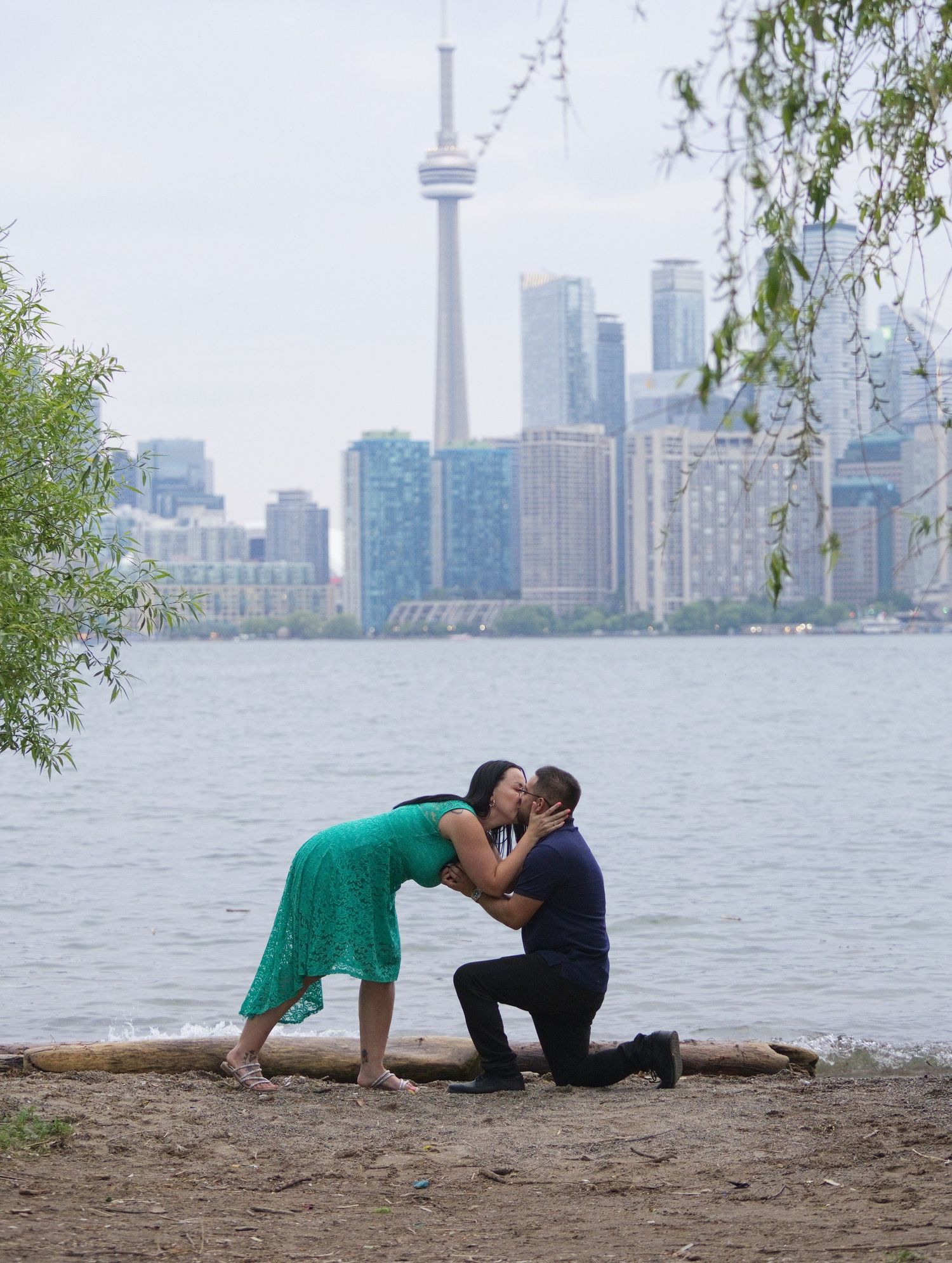 Couple shares a romantic proposal kiss on Toronto waterfront with CN Tower skyline backdrop.