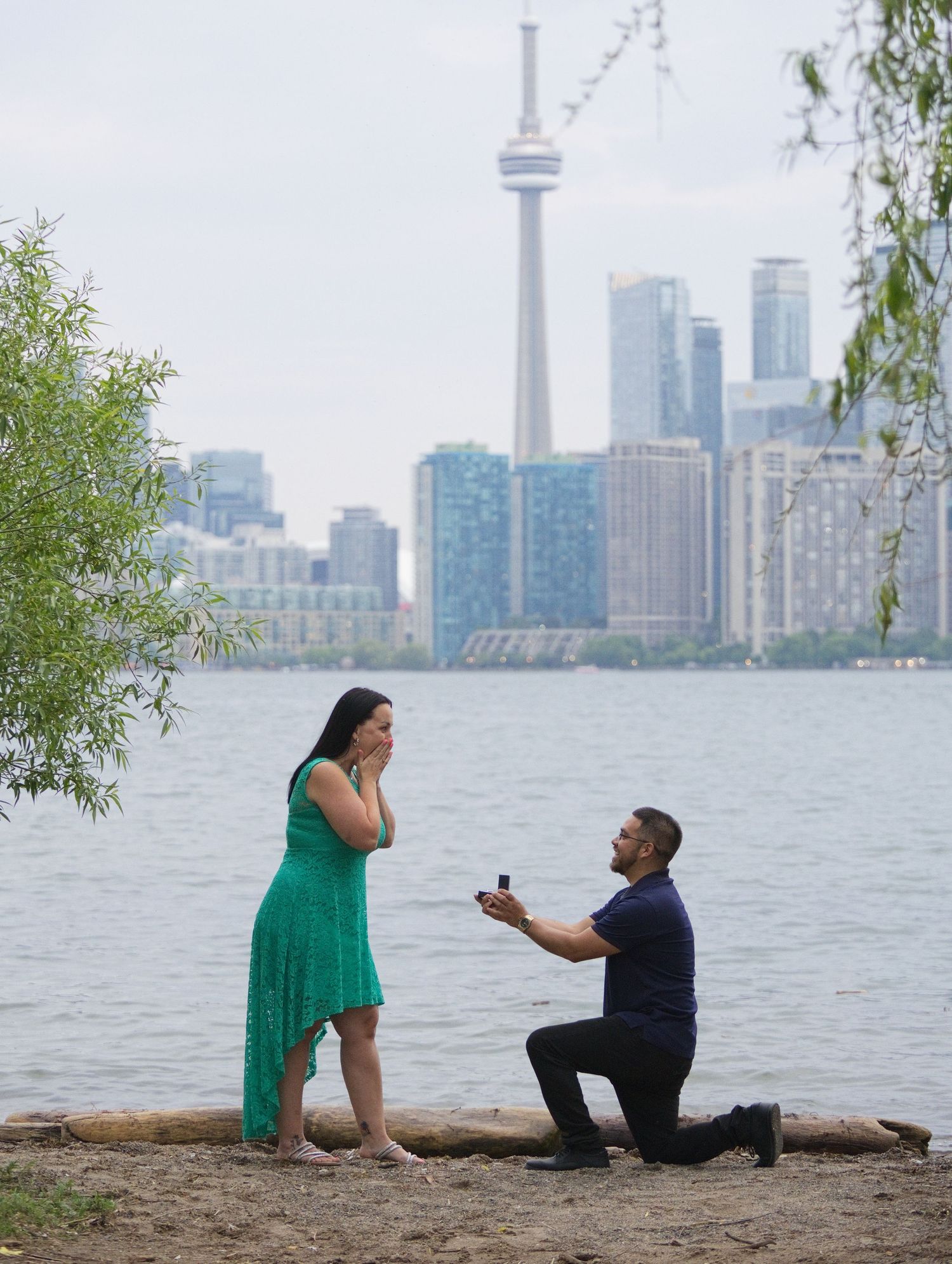 Man proposes on one knee to woman in green dress by Toronto waterfront with CN Tower skyline backdrop.