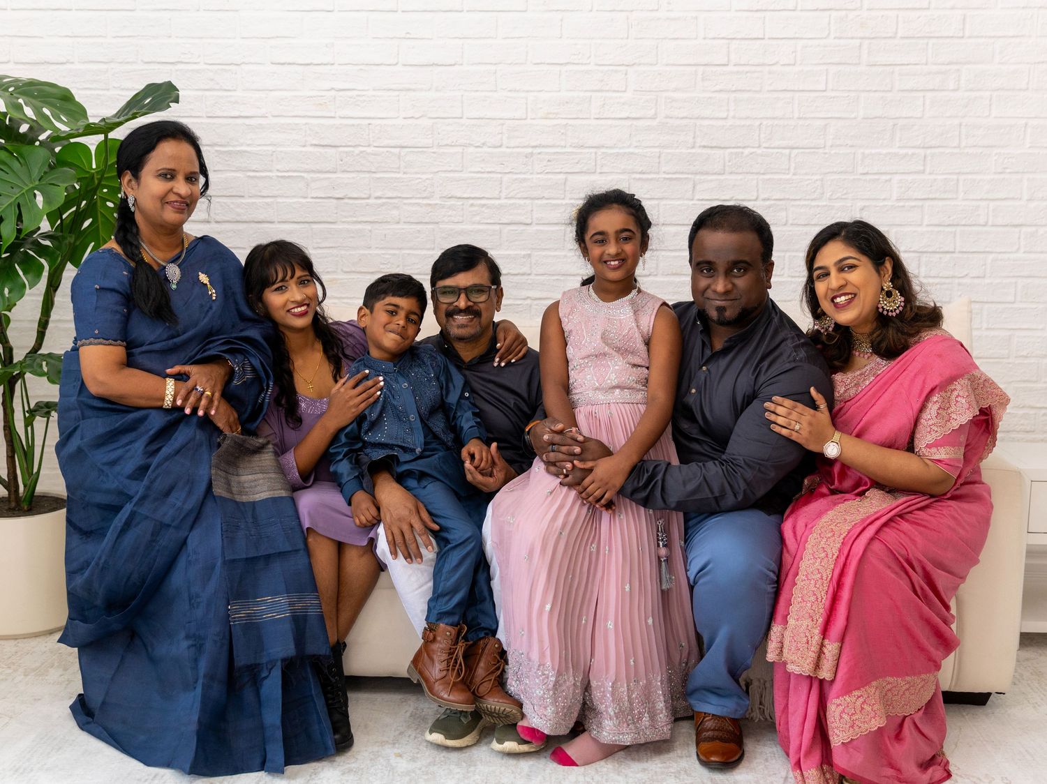 A joyful South Asian family of seven poses together on a white couch, dressed in elegant blue and pink traditional attire.