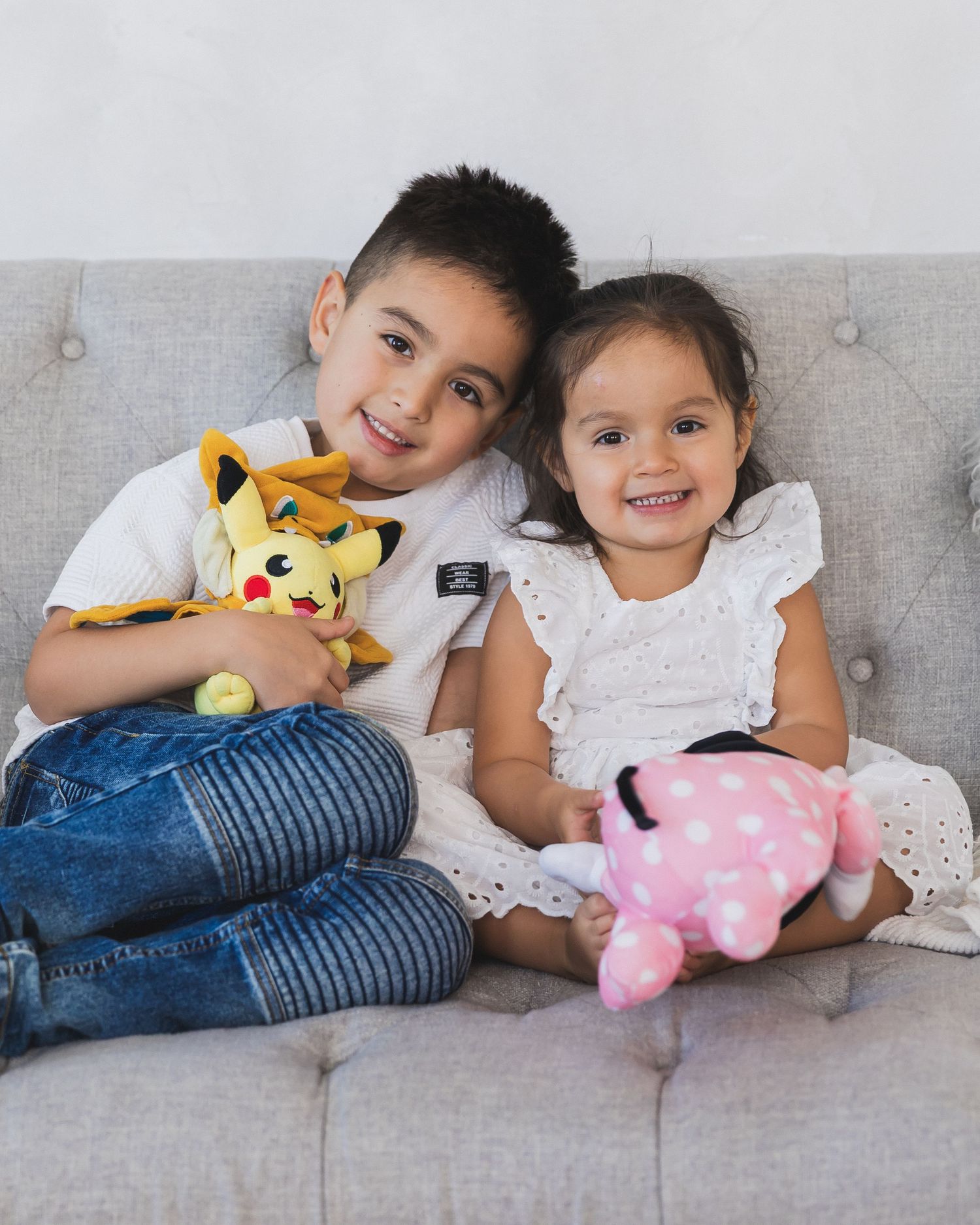 Brother and sister sitting on gray couch holding Pikachu and pink stuffed animal toys.