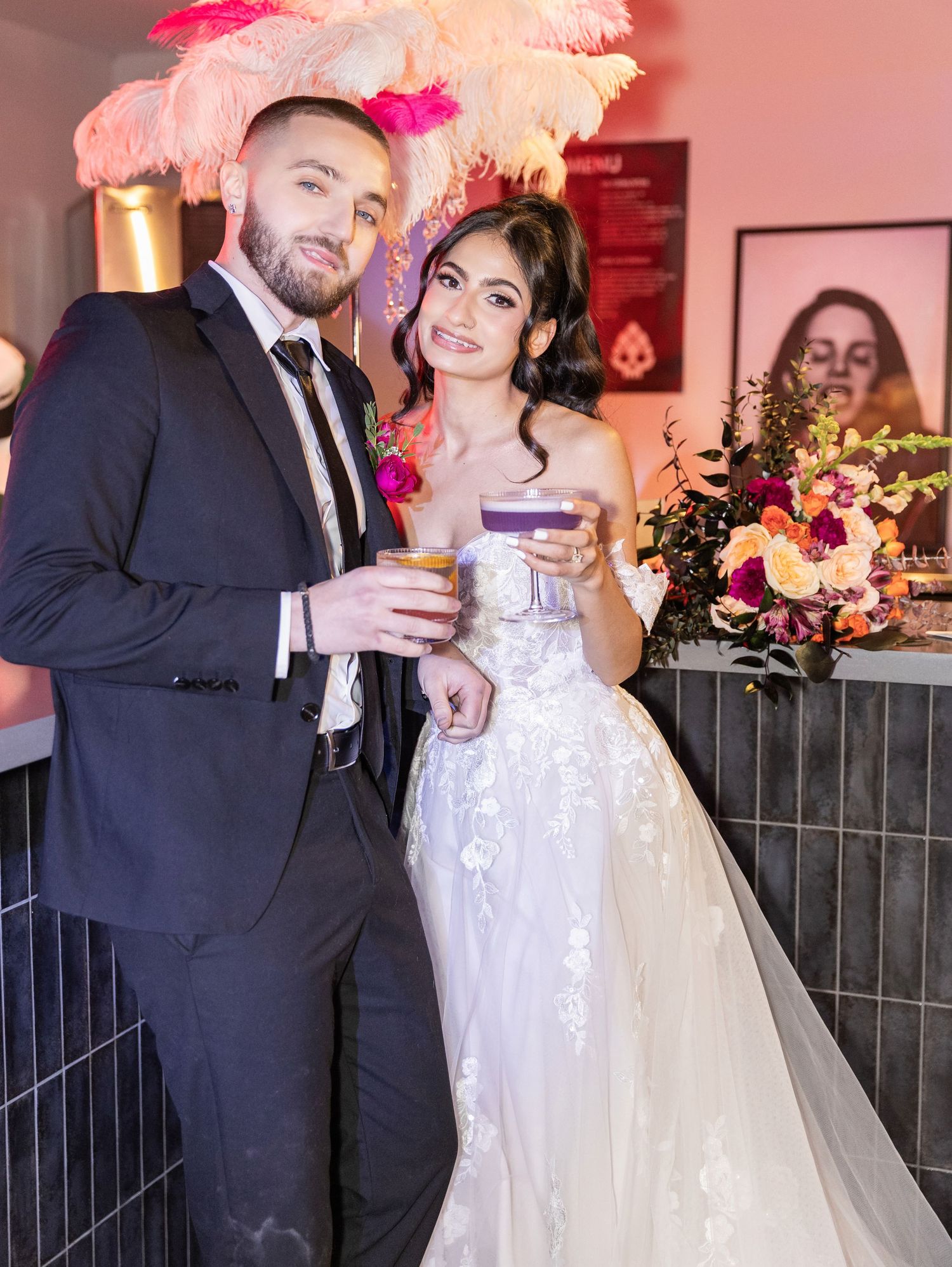 Bride and groom holding cocktails at bar with pink feather decor and colorful wedding bouquet.