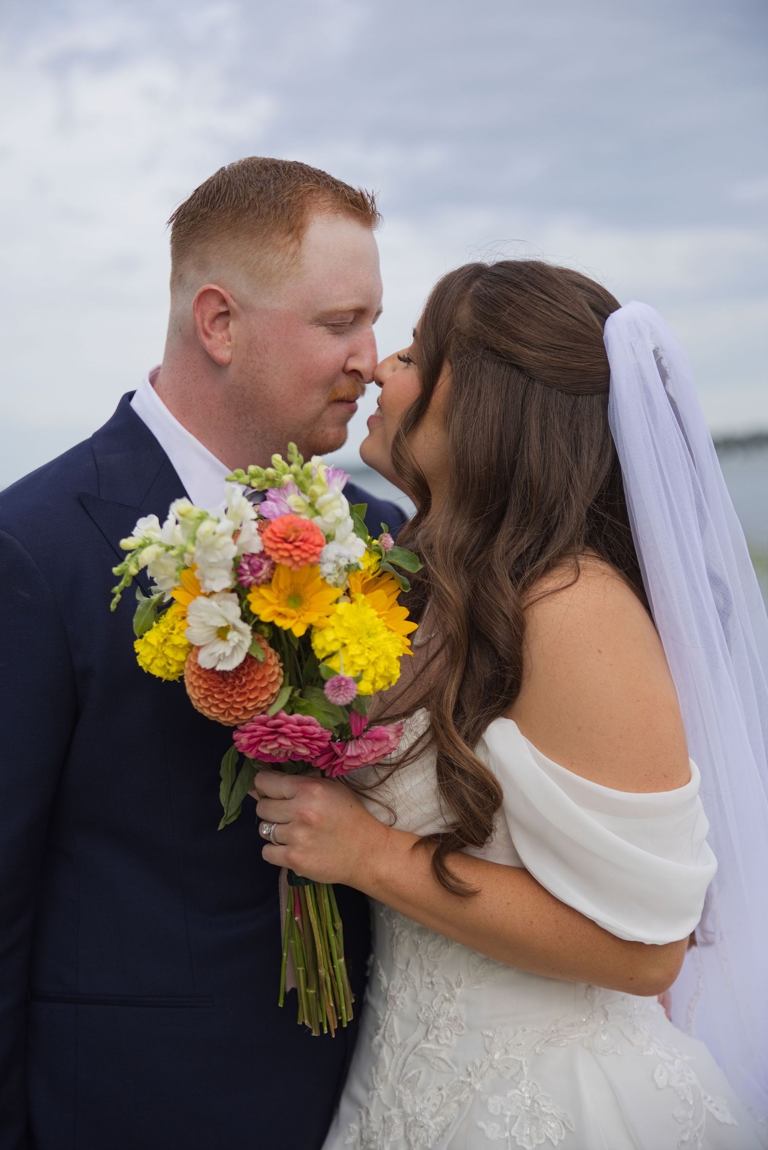 Bride and groom sharing a romantic nose-to-nose moment holding a colorful bouquet of yellow, pink, and white flowers.