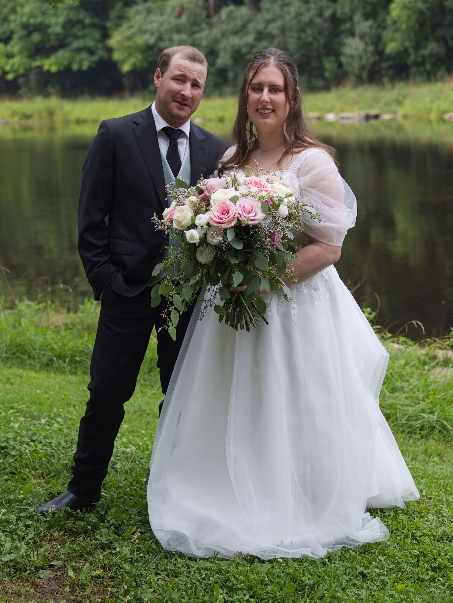 Bride in white off-shoulder gown holding pink bouquet poses with groom in black suit by a pond.