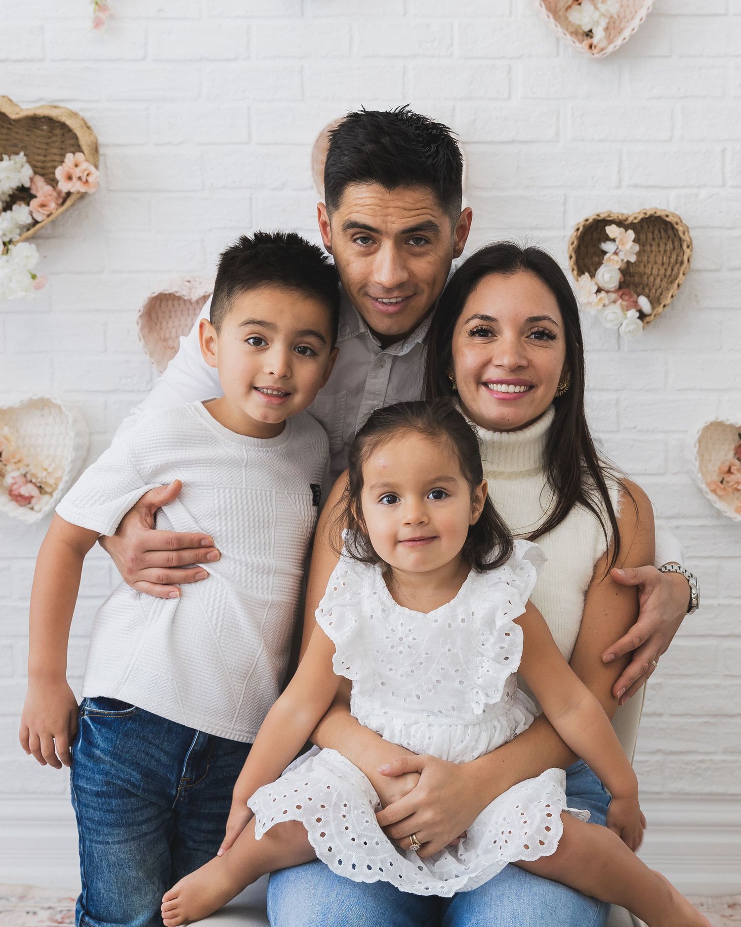 Family of four portrait with parents and two young children posing in front of a white wall with heart decorations.