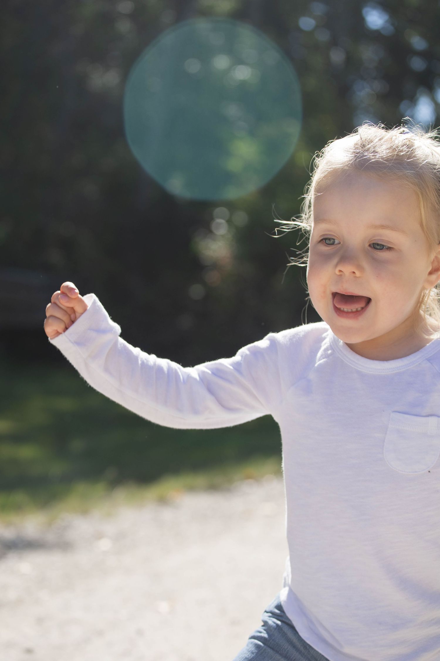 A joyful toddler girl in a white long-sleeve shirt runs outdoors with arm raised, sunlight creating a lens flare.