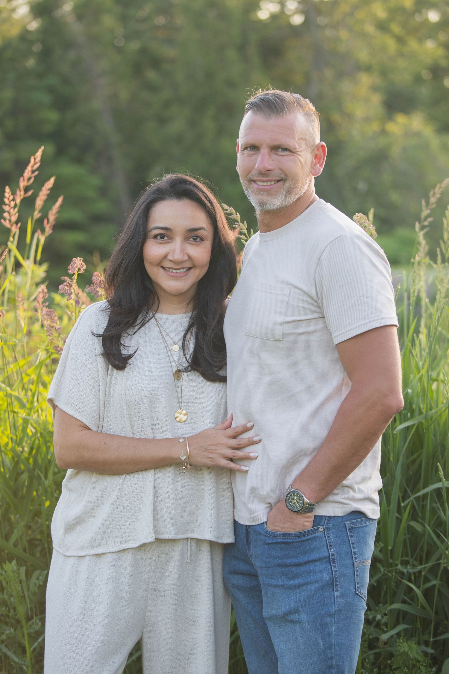 Smiling middle-aged couple posing together in golden hour sunlight among tall green grasses.