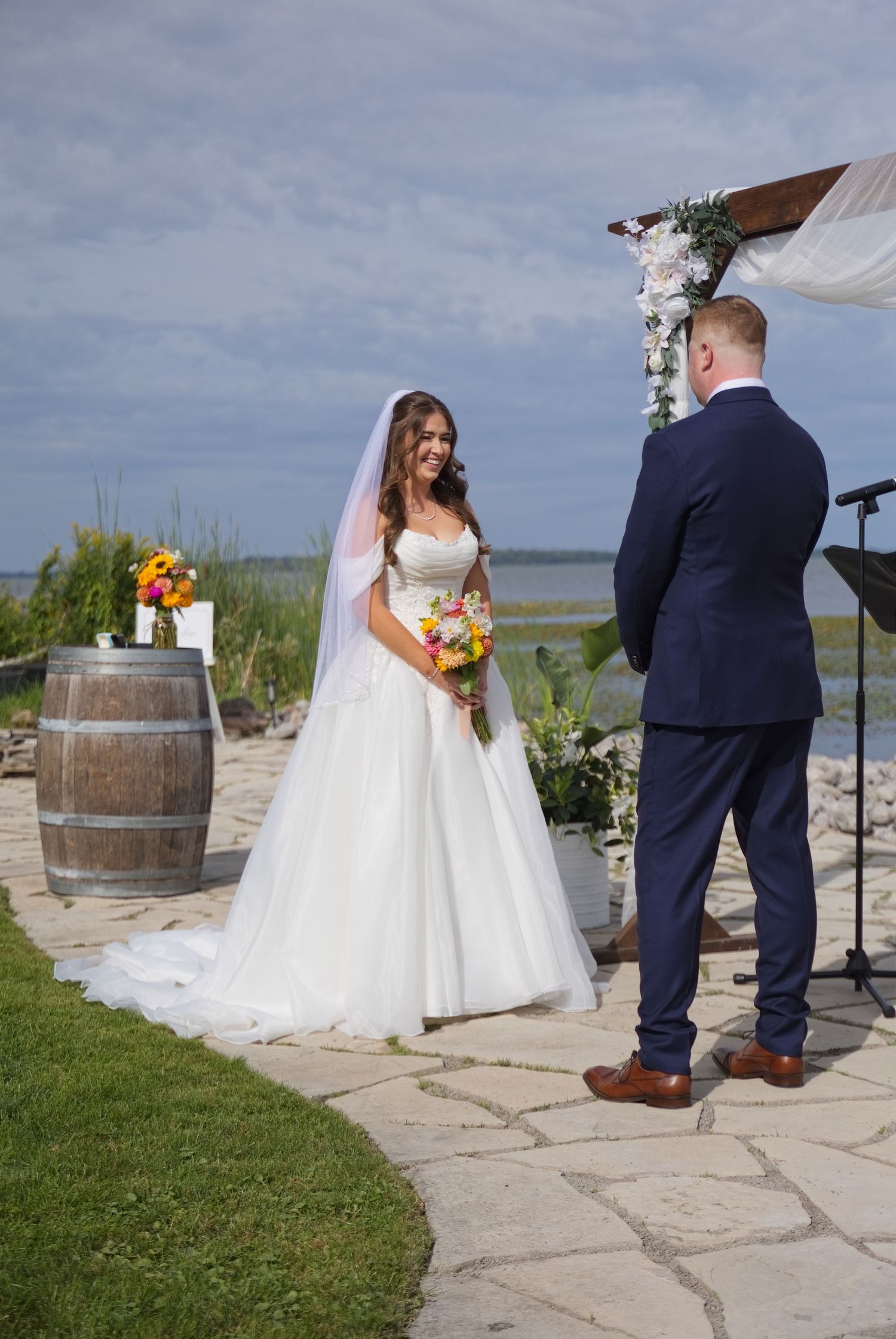 Bride in white gown smiling at groom during outdoor waterfront wedding ceremony under floral arch.