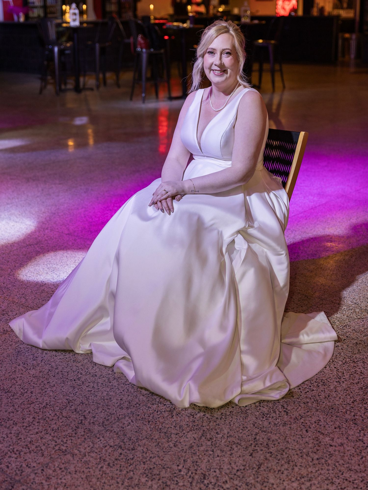 Blonde bride in elegant white satin ball gown seated on gold chair with pink venue lighting.