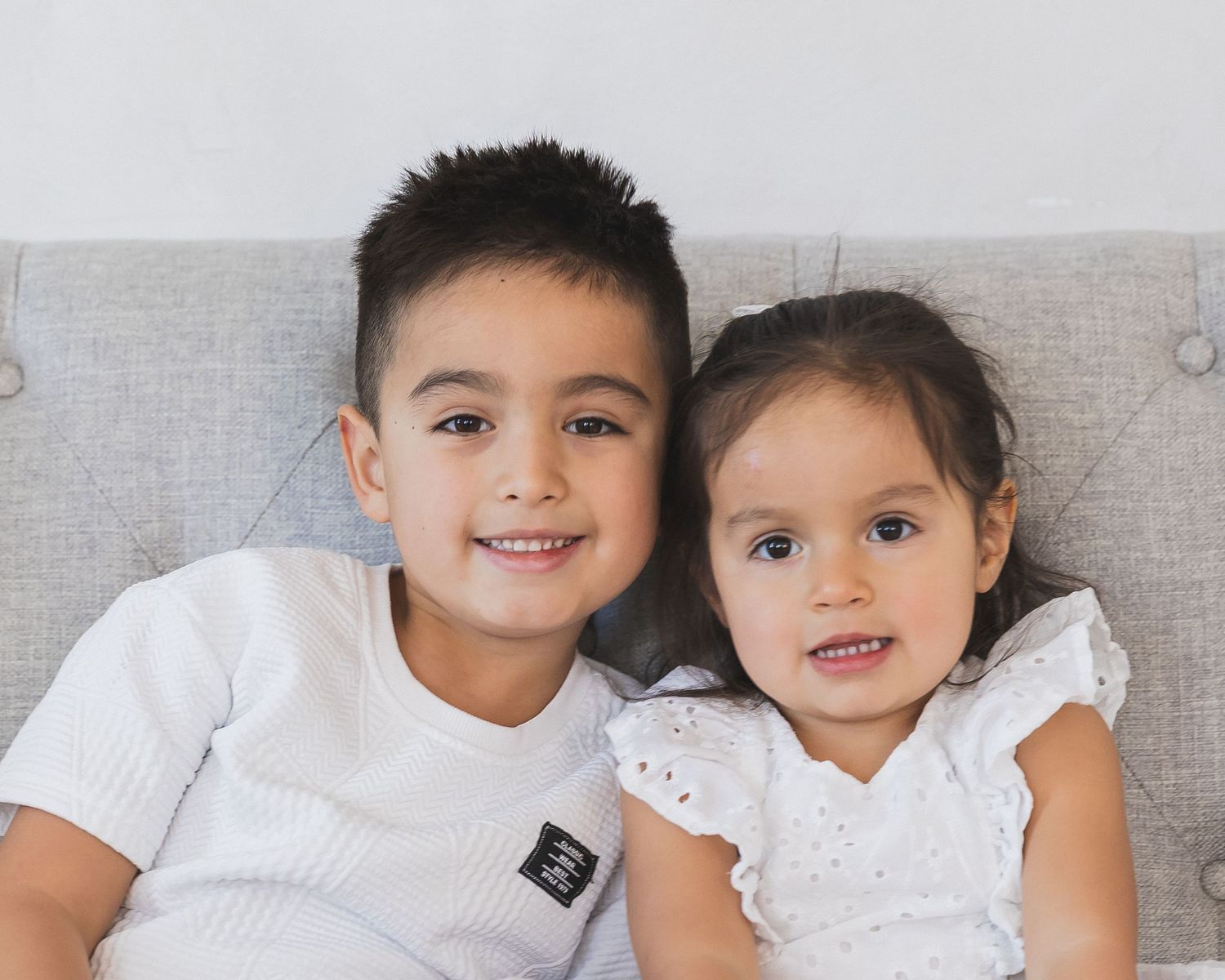 Two young siblings in white outfits smiling together on a gray couch.