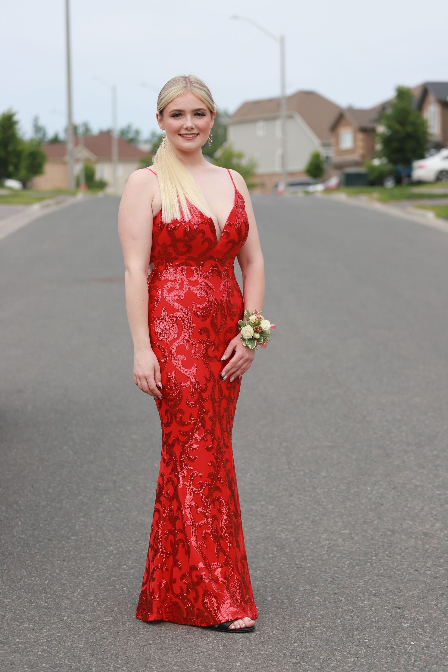Young woman in a red sequin mermaid prom gown with wrist corsage posing on a suburban street.