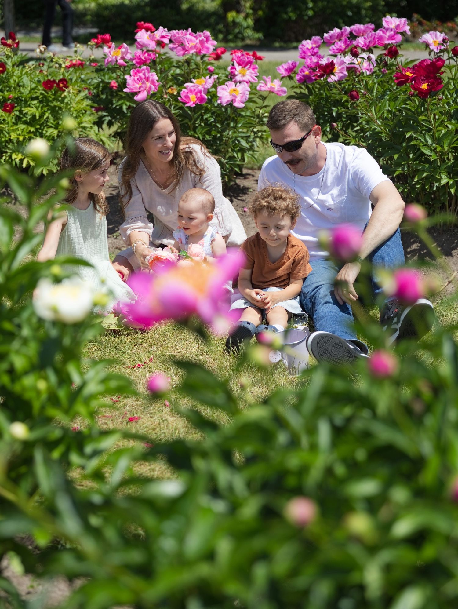 Family sitting together in a blooming garden surrounded by vibrant pink and red peony flowers on a sunny day.