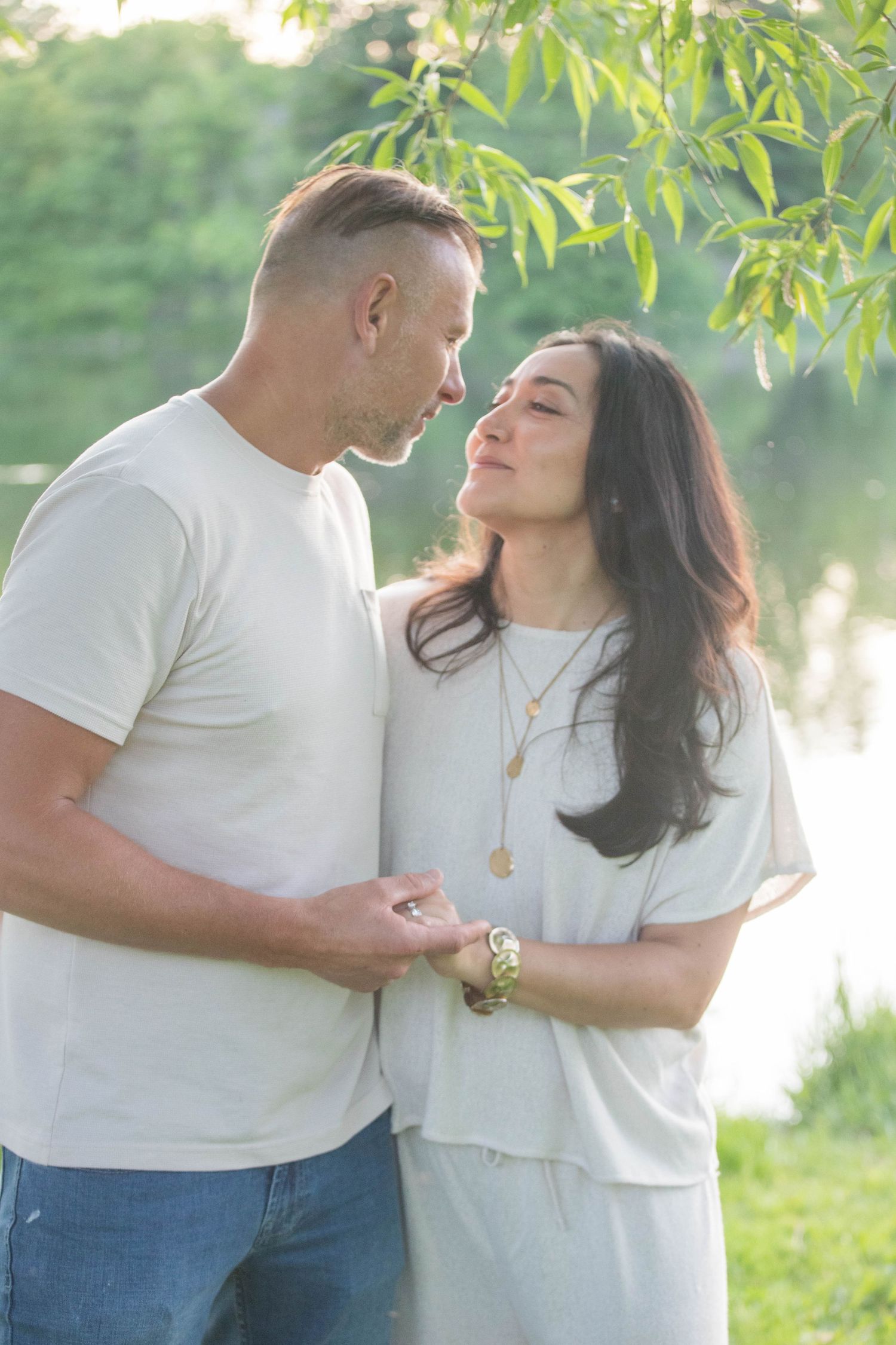 Romantic couple sharing tender moment near sunlit pond under willow branches.
