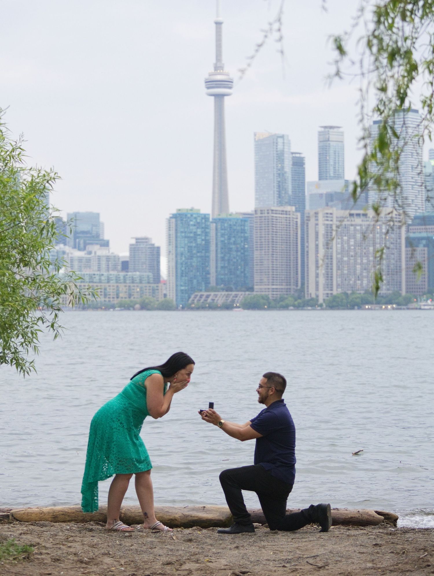 Man proposes on one knee to woman on Toronto waterfront beach with CN Tower skyline backdrop.