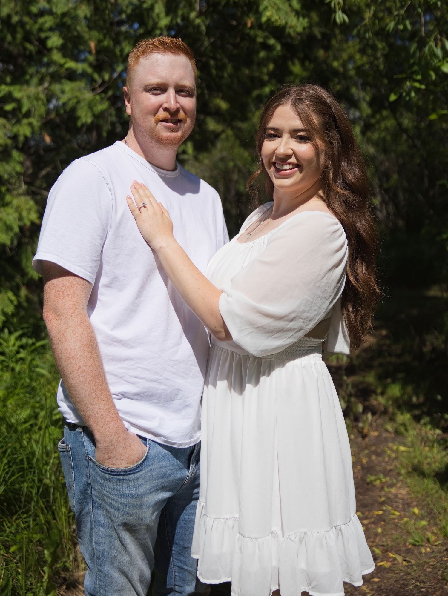 A smiling couple poses outdoors on a garden path, woman in white dress with hand on mans chest.