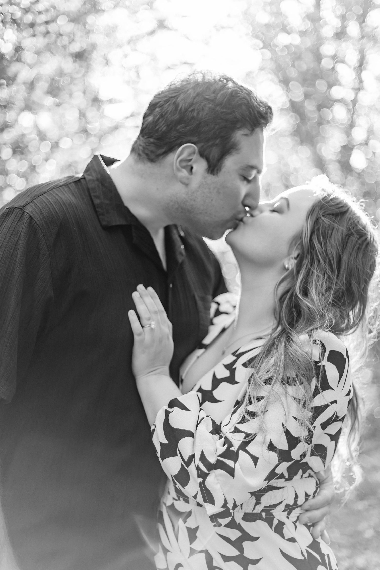 Black and white romantic photo of couple sharing a kiss outdoors in soft light.