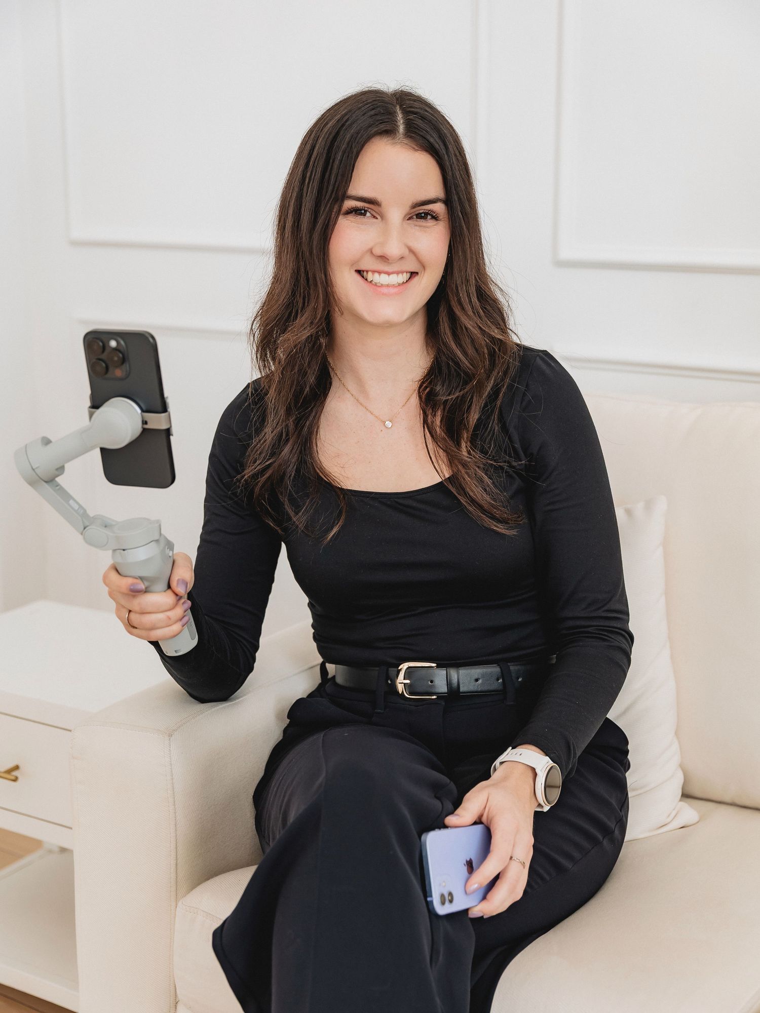Woman in black outfit holds a smartphone gimbal stabilizer while sitting on a white couch, smiling confidently.