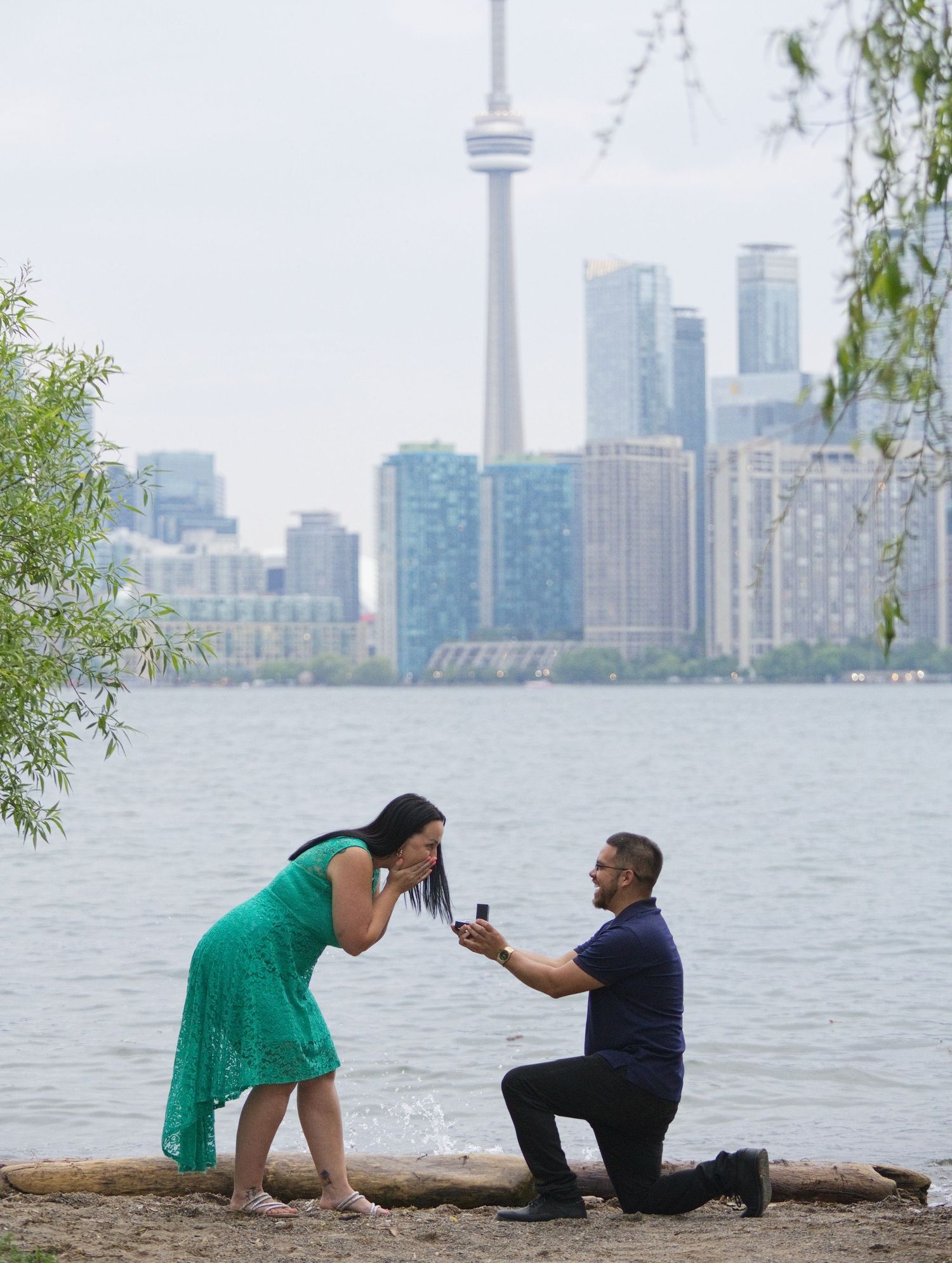 Man proposes to woman on one knee at Toronto waterfront with CN Tower skyline backdrop.