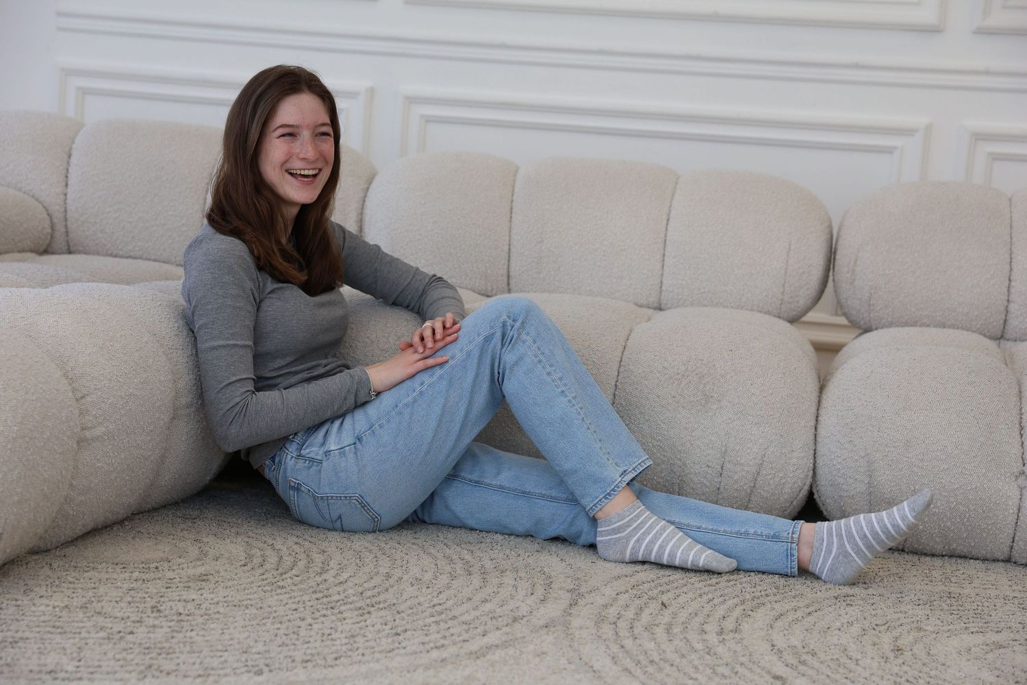 Young woman sitting on floor laughing beside a large beige sectional sofa, wearing jeans and striped socks.