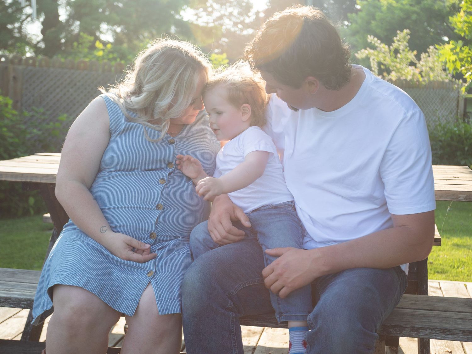 Pregnant woman and man sitting on bench with toddler in sunny garden setting.