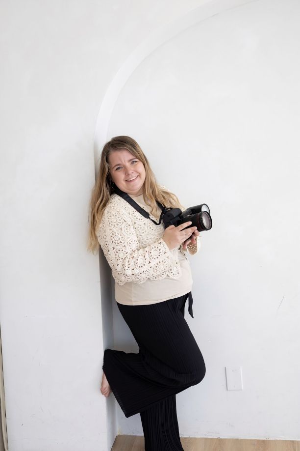 Smiling woman with long blonde hair holding a DSLR camera, wearing a cream sweater and black pants, leaning against a white wall.