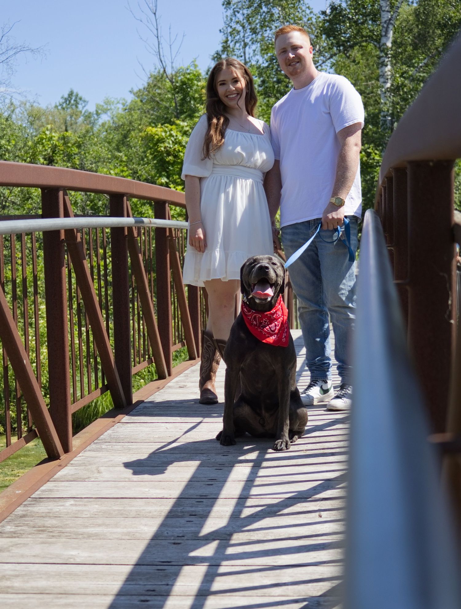 Couple in white dress and white t-shirt standing on wooden bridge with black Labrador wearing red bandana.