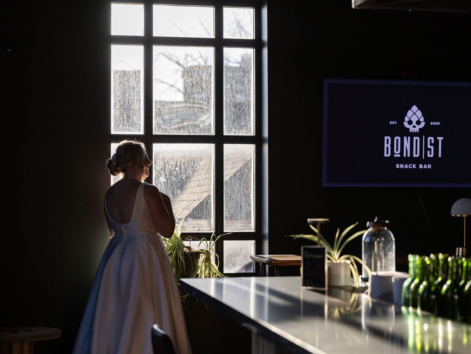 Bride in white gown stands by large window at Bond St Snack Bar venue counter.