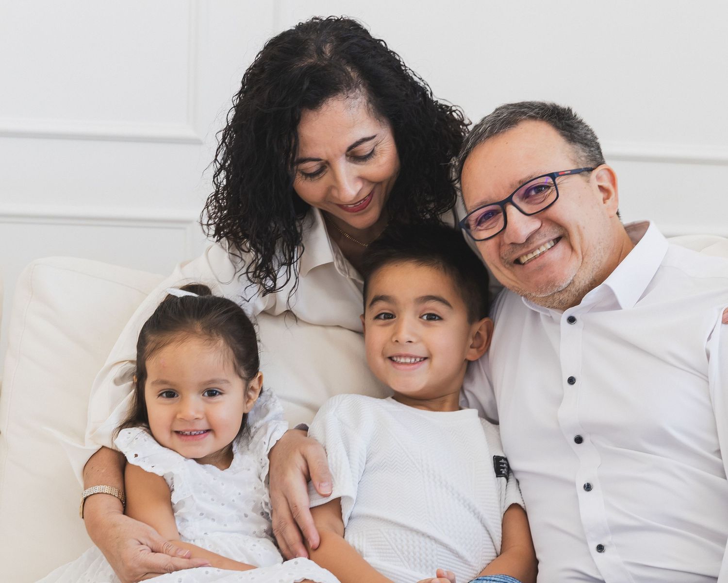 Smiling parents with two young children cuddling together on a white couch indoors.