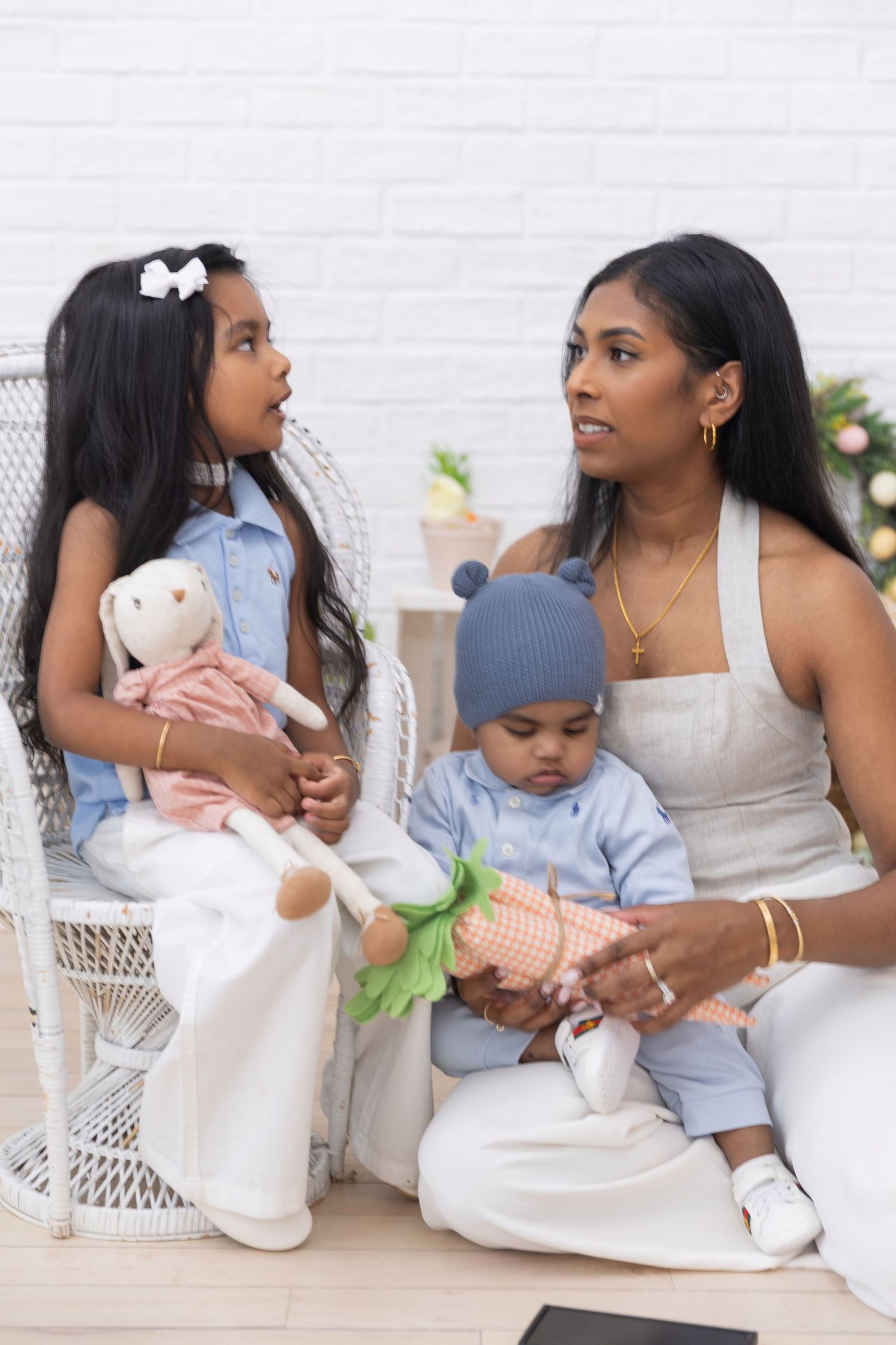 Mother sitting with young daughter holding a bunny doll and baby boy in Easter-themed photo studio.