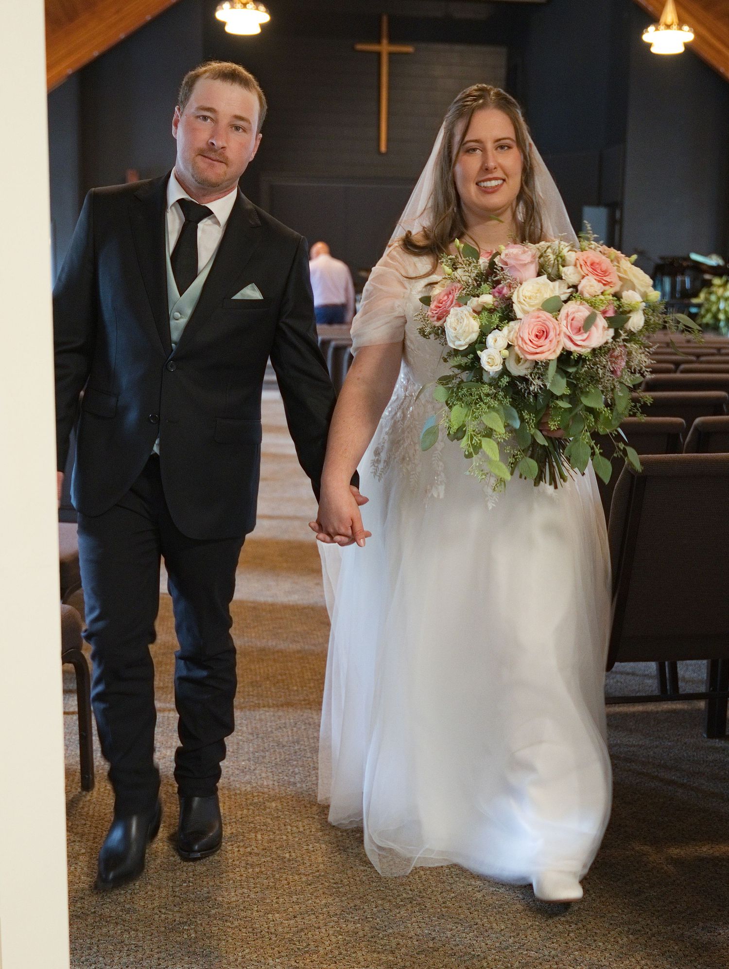 Bride and groom walking hand in hand down church aisle, bride holding pink and white rose bouquet in wedding dress.