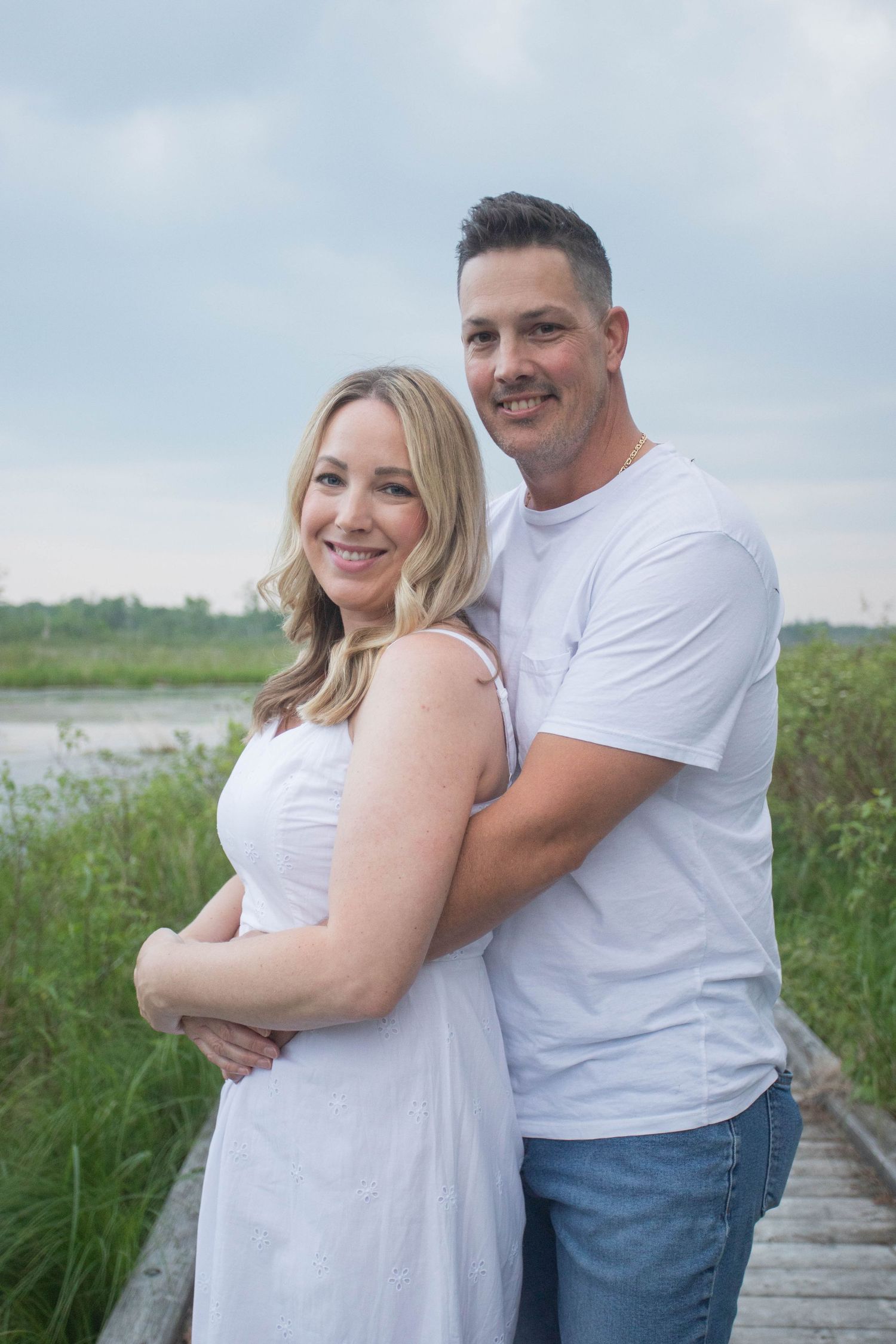 A man in a white t-shirt embraces a woman in a white dress on a wooden boardwalk.