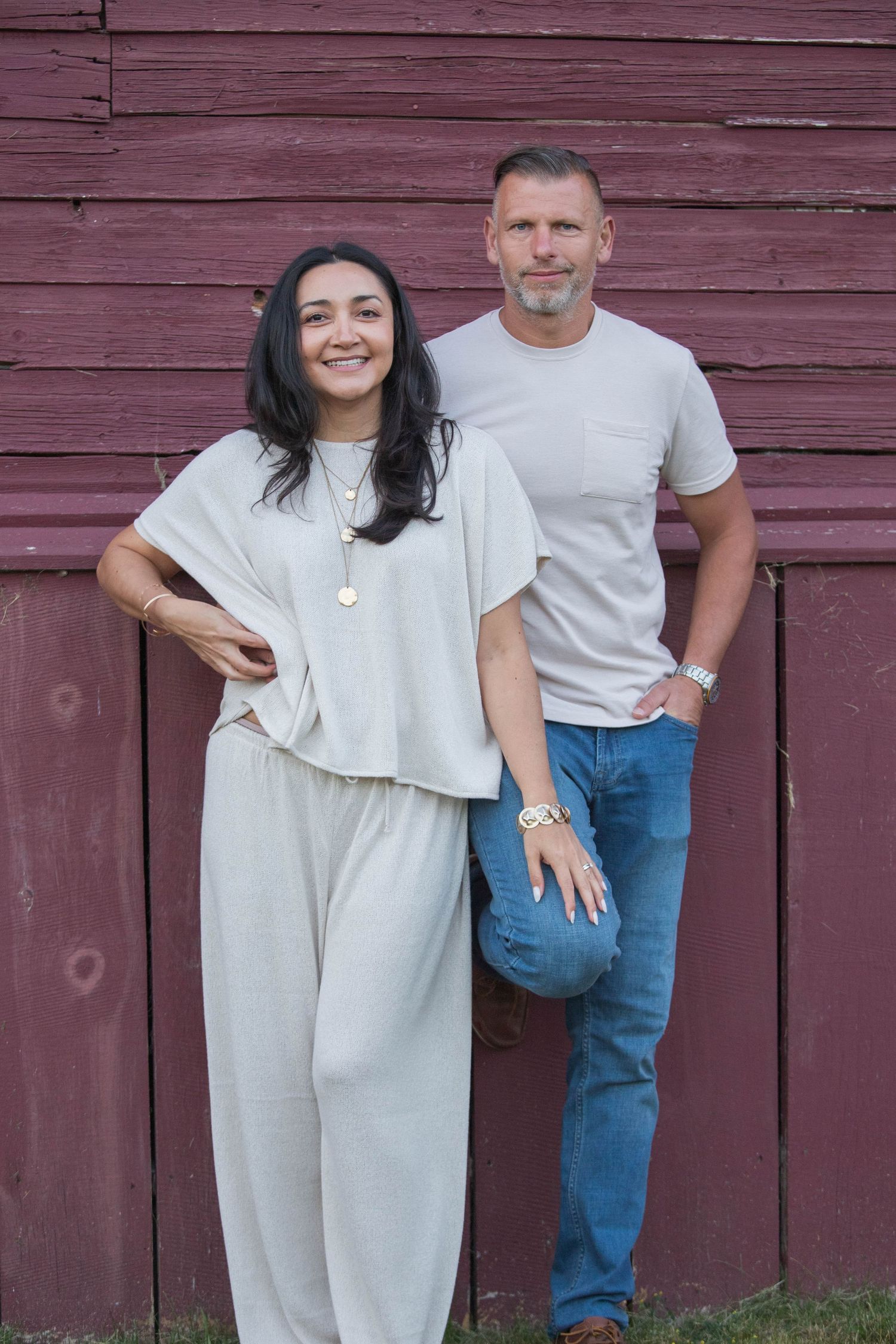 Stylish couple posing casually against a rustic red barn wall, dressed in coordinating neutral tones.