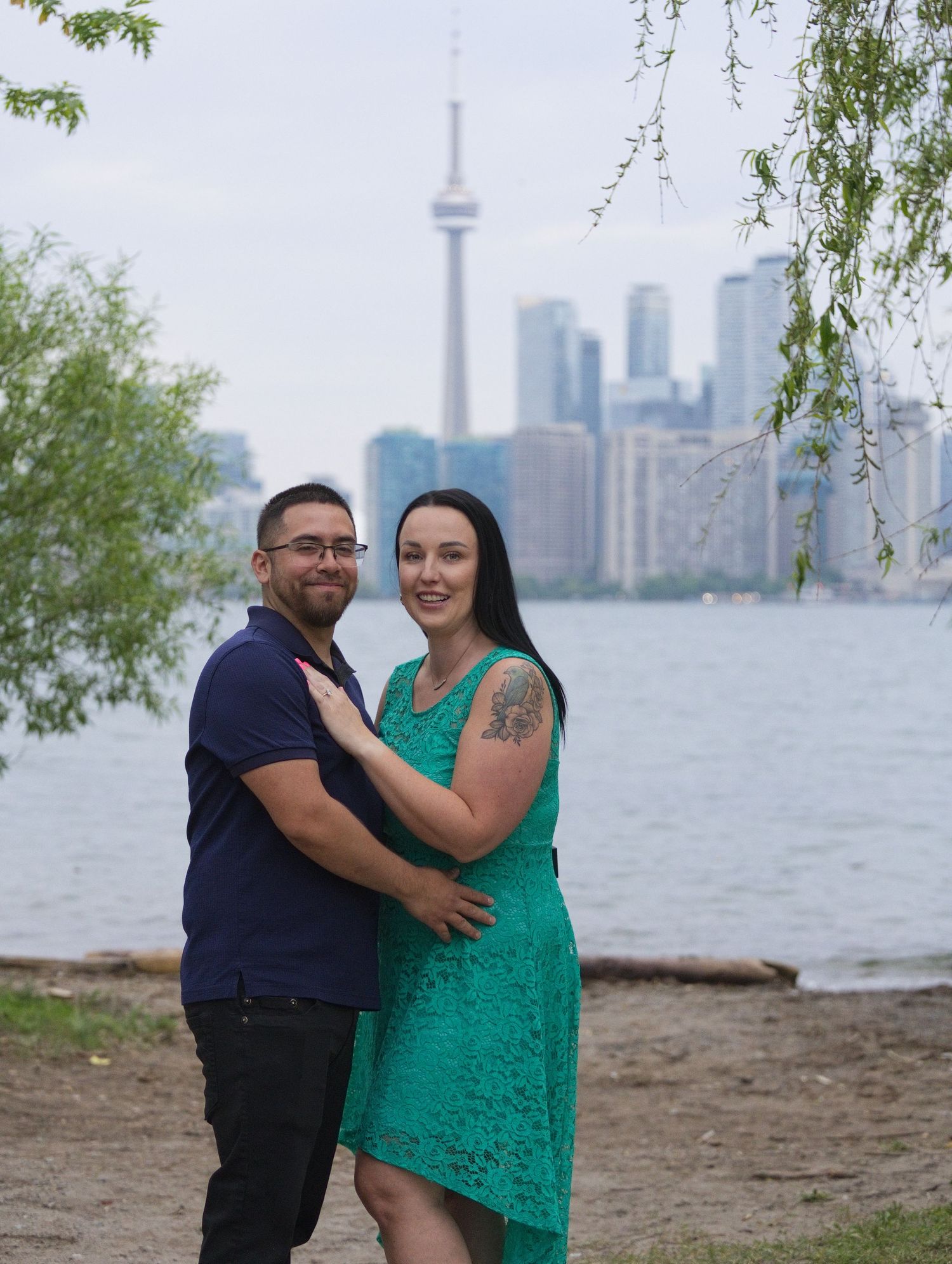 Couple embracing on Toronto Island waterfront with CN Tower and city skyline visible in the background.