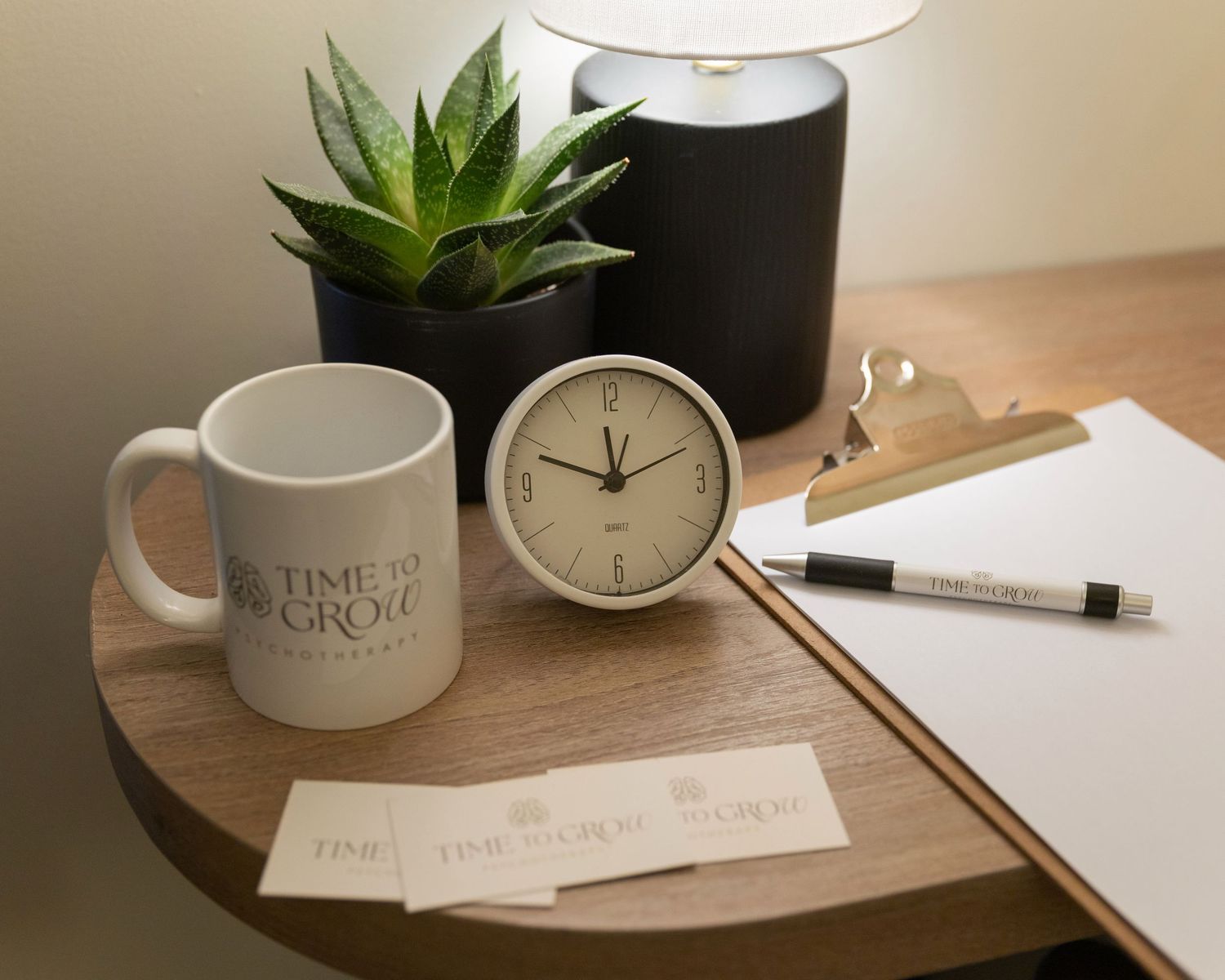A wooden desk with a Time to Grow mug, clock, plant, clipboard, pen, and branded cards in a cozy office setting.