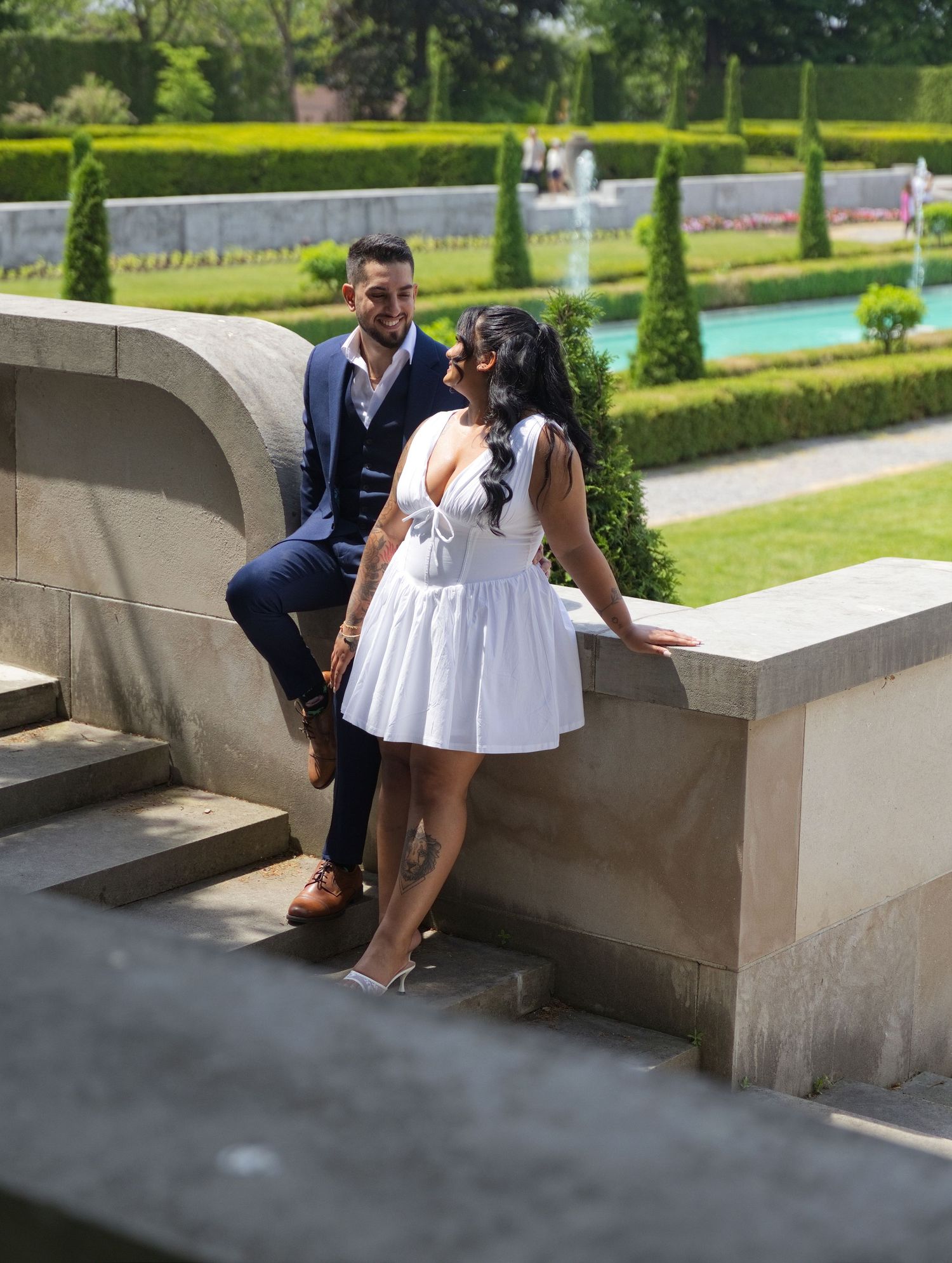 Couple in formal wear sitting on stone steps in a manicured garden, woman in white dress, man in navy suit.