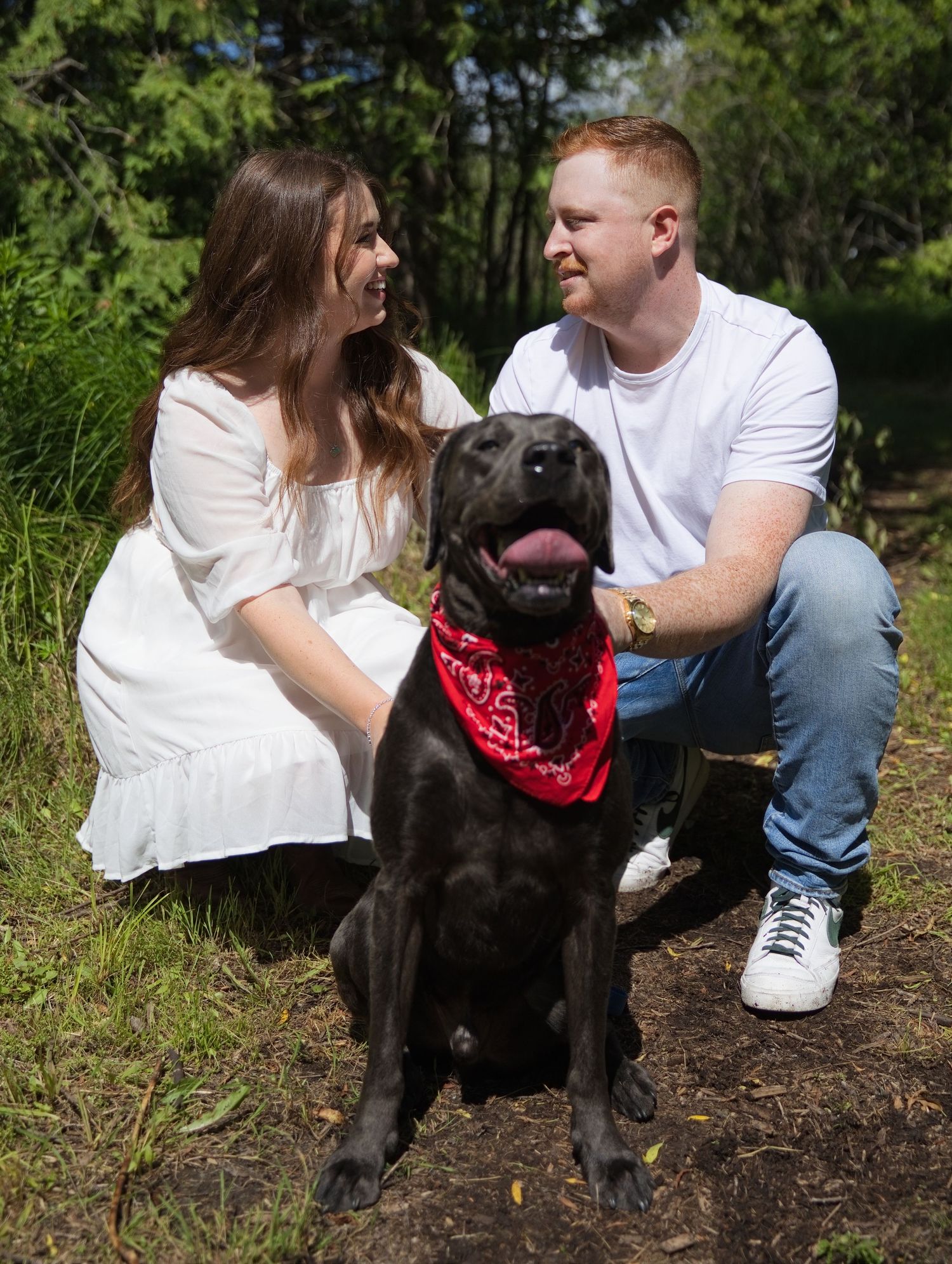 Couple in white outfits posing outdoors with a black Labrador wearing a red bandana in a sunny forest setting.