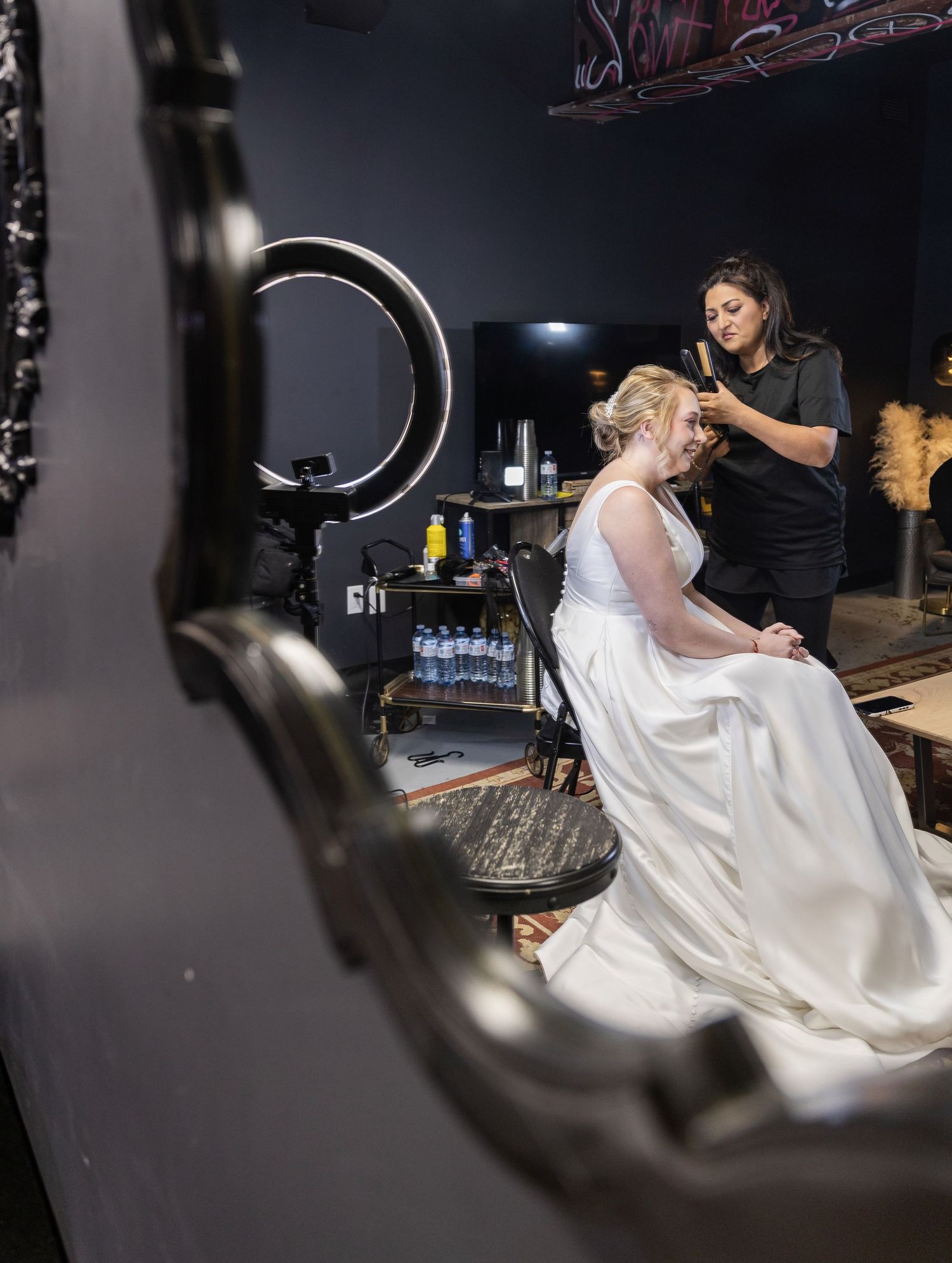Bride in white wedding gown getting hair styled by makeup artist with ring light.