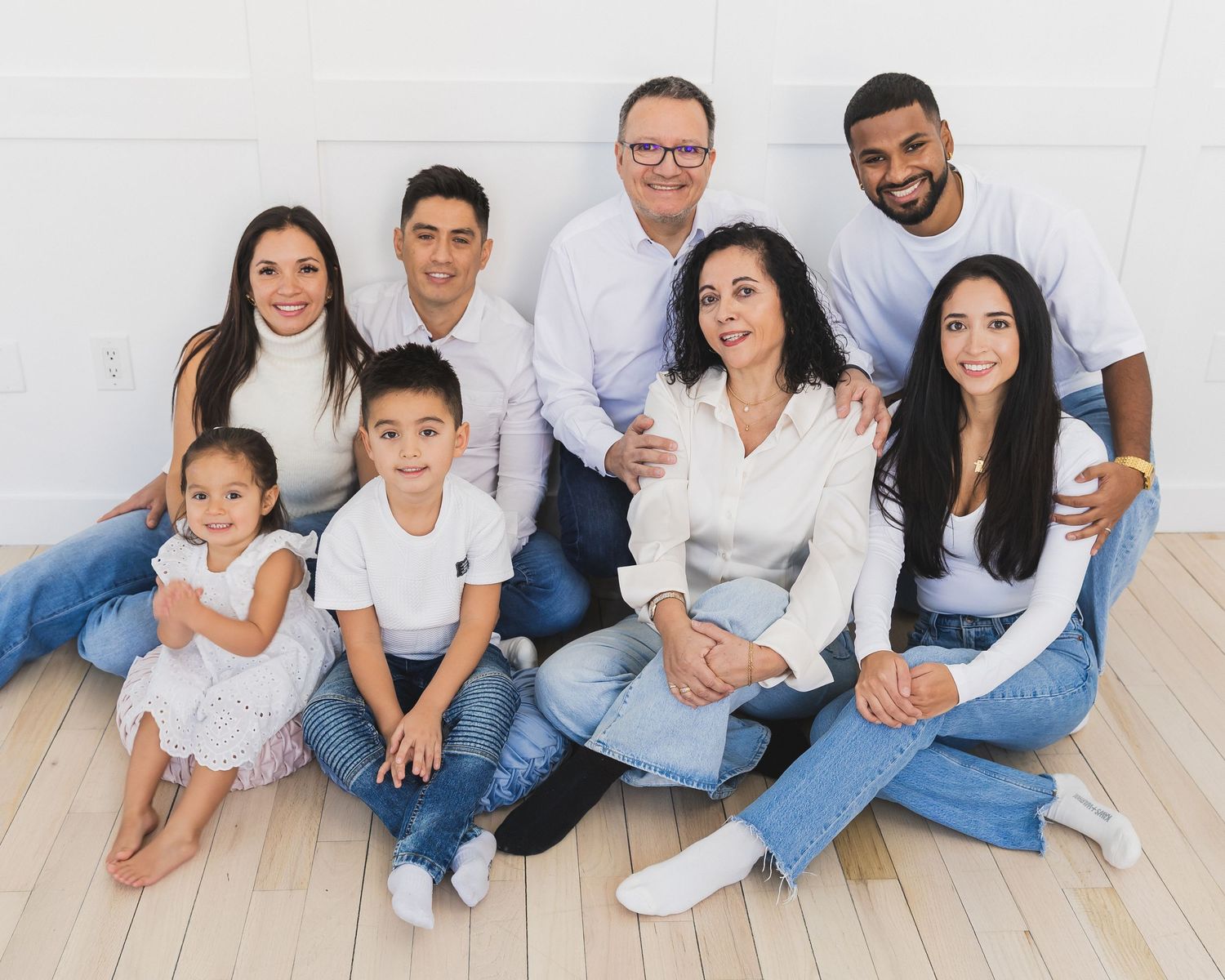 Extended family of nine posing together on a light wood floor, dressed in white tops and blue jeans for a portrait.