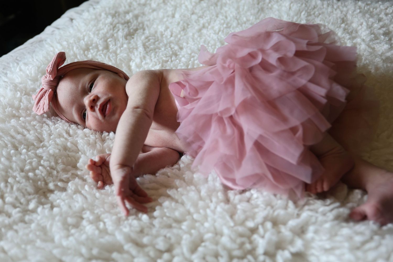 A newborn baby girl in a pink tutu skirt and floral headband lies on a fluffy white blanket.