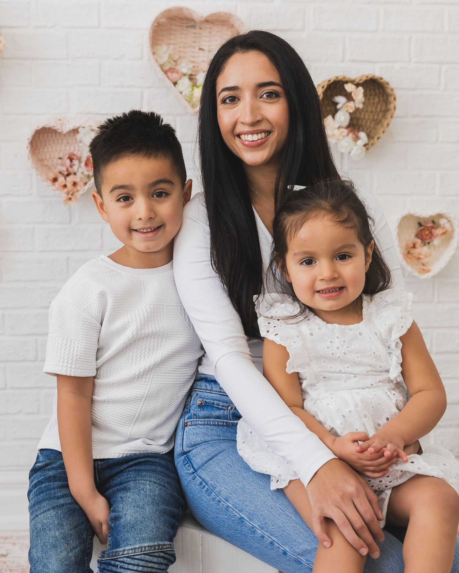 Young mother smiling with her two children, a boy and a toddler girl, in front of a heart-decorated white wall.