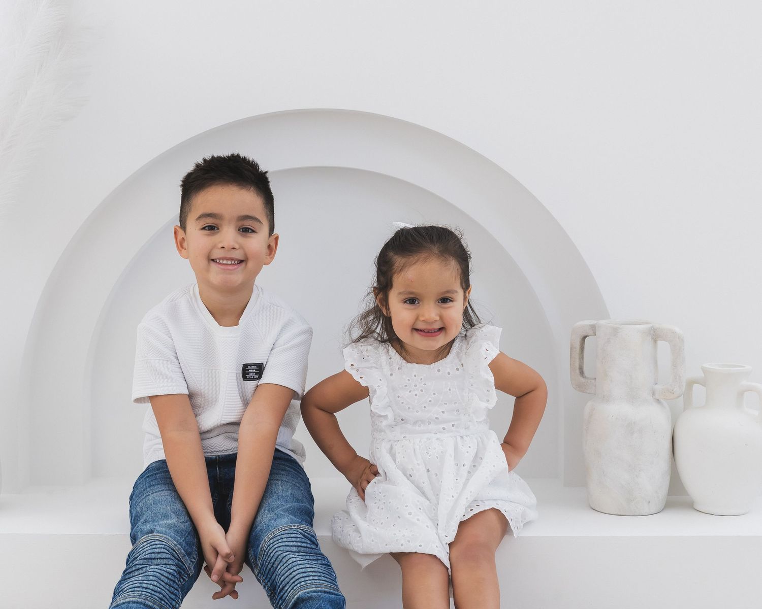 Two young Hispanic siblings in white outfits smile while sitting together in front of a white arch backdrop.