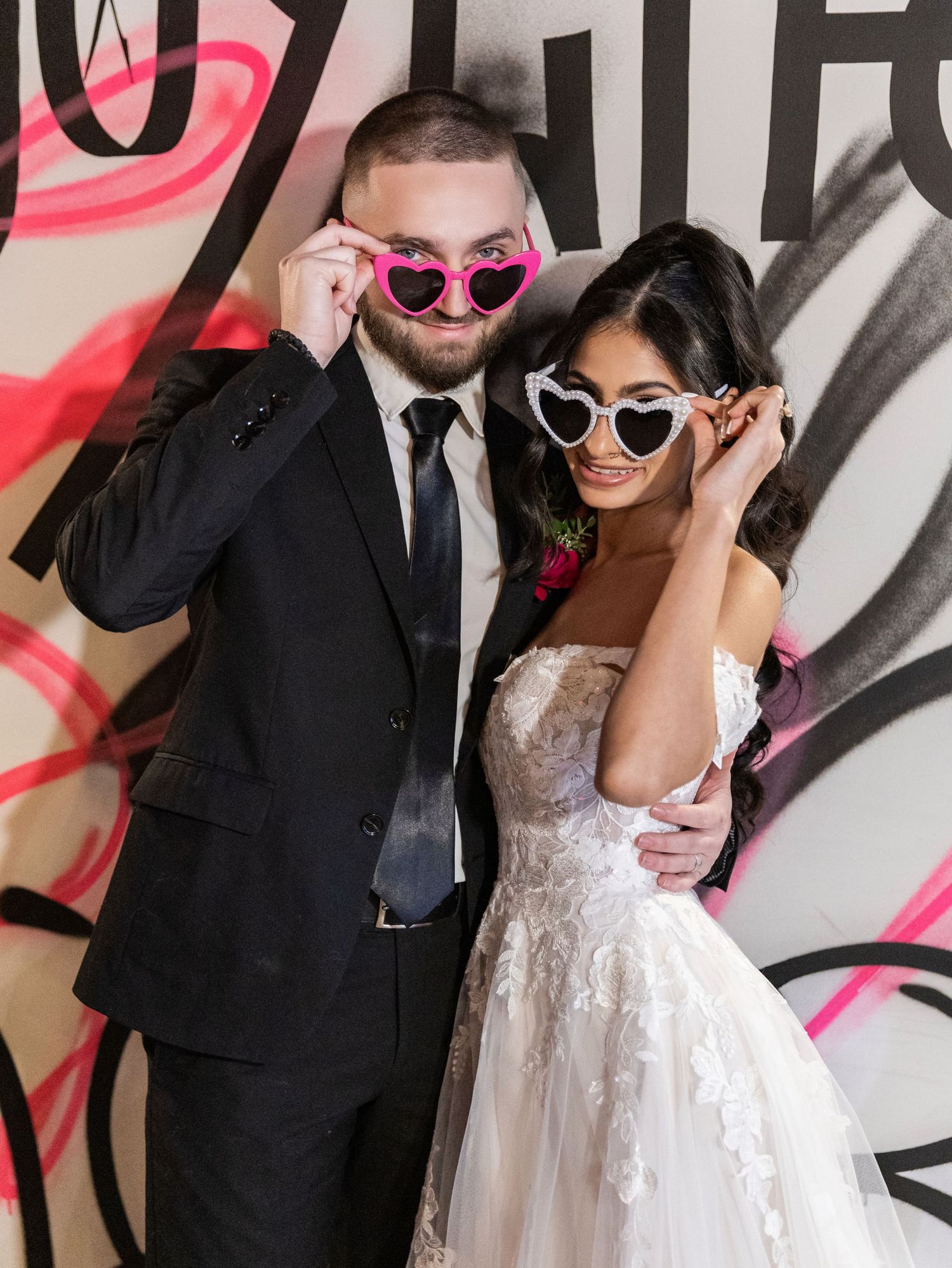 Bride and groom posing playfully with heart-shaped sunglasses in front of a colorful graffiti backdrop.