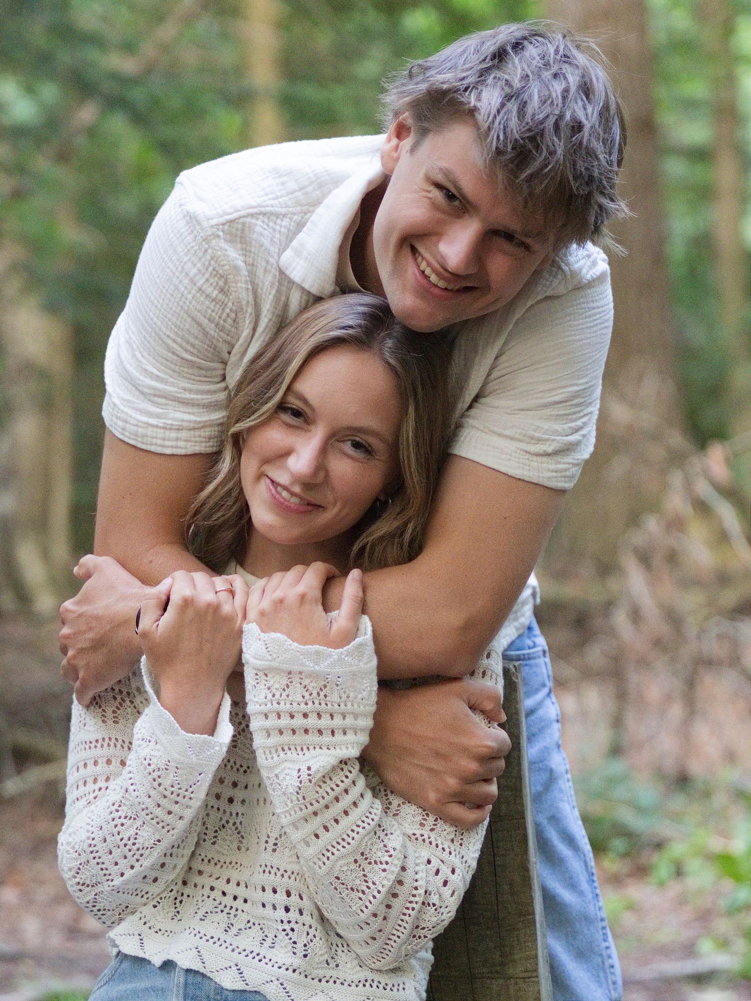 A young couple embracing warmly in a forest, the man hugging the woman from behind, both smiling happily.