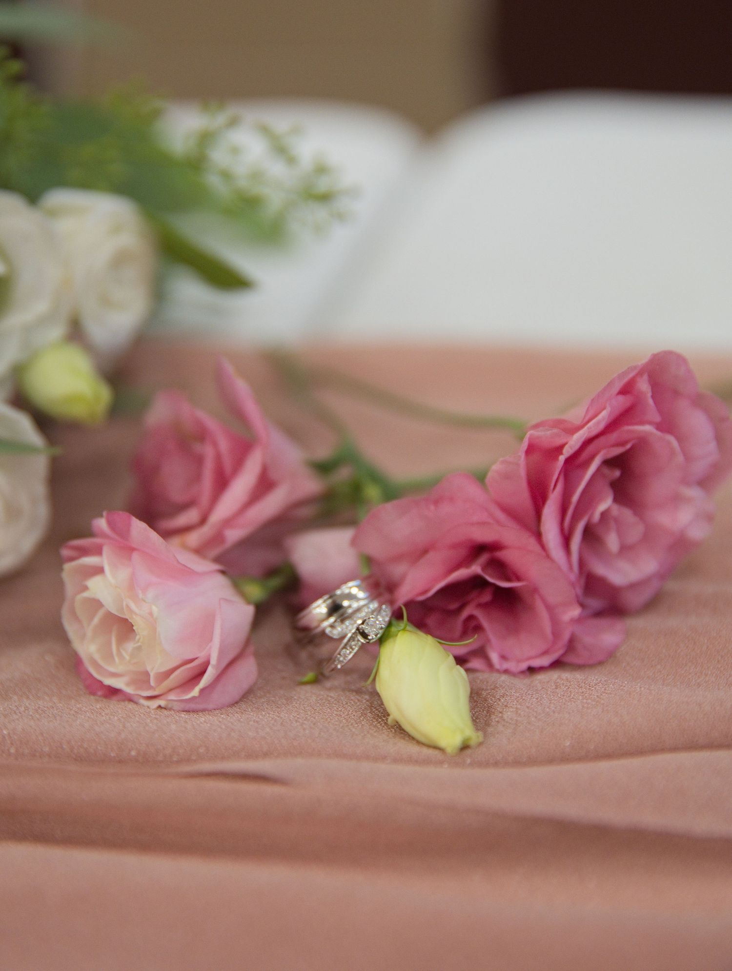 Pink roses and wedding rings on a blush pink tablecloth with white flowers in the background.