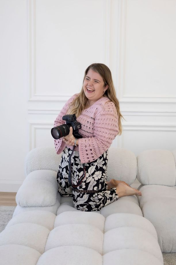 A smiling woman kneeling on a white tufted sofa, holding a DSLR camera, wearing a pink cardigan and floral skirt.