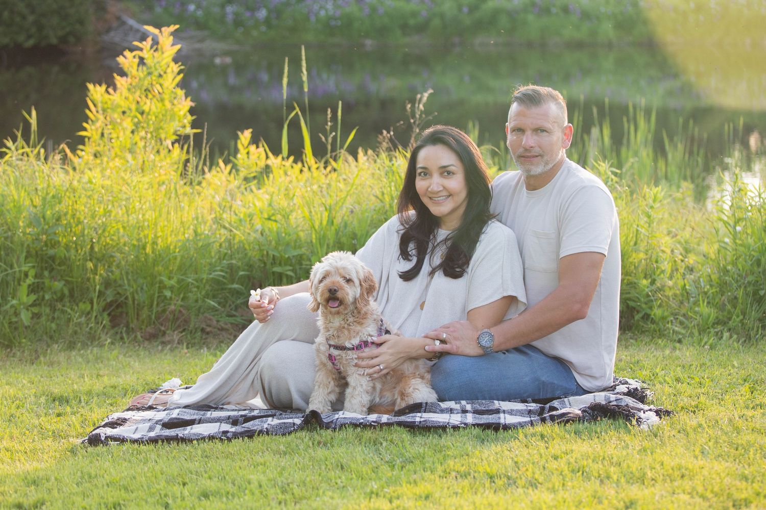 Couple sitting on plaid blanket outdoors with fluffy dog, golden hour lighting near pond with wildflowers.