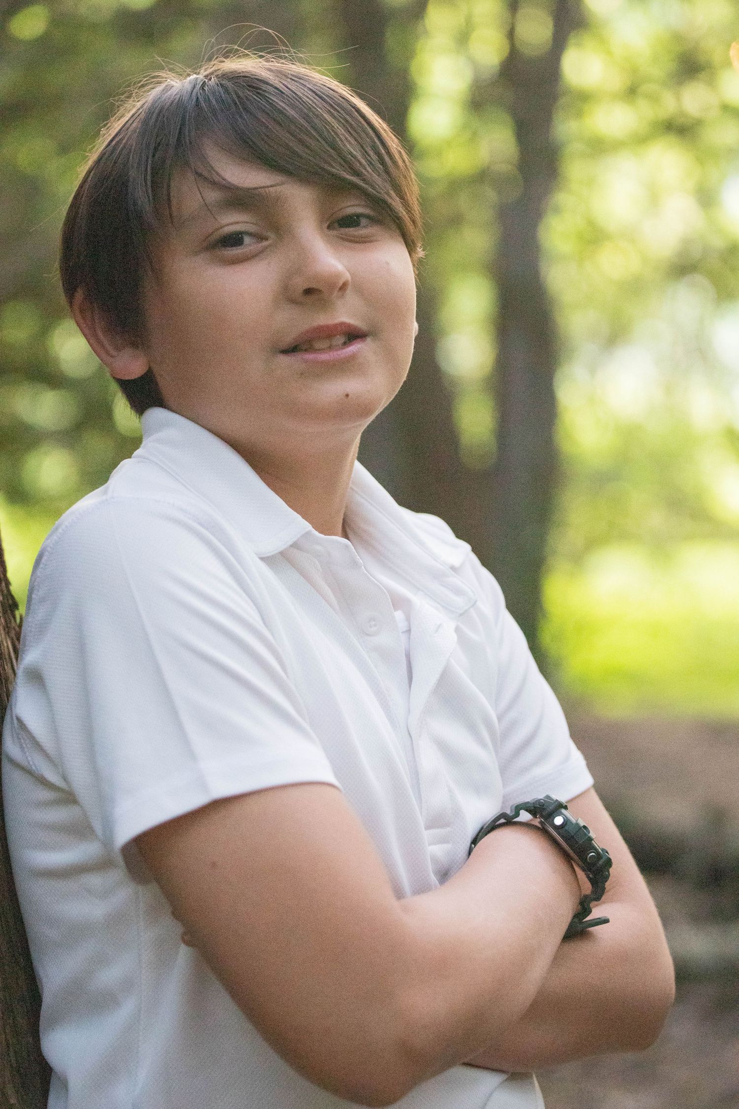 Smiling preteen boy in white shirt with arms crossed outdoors.