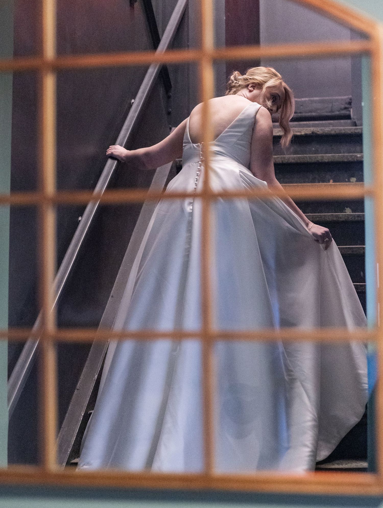 Bride in elegant white ball gown ascending dark staircase, viewed through window frame.