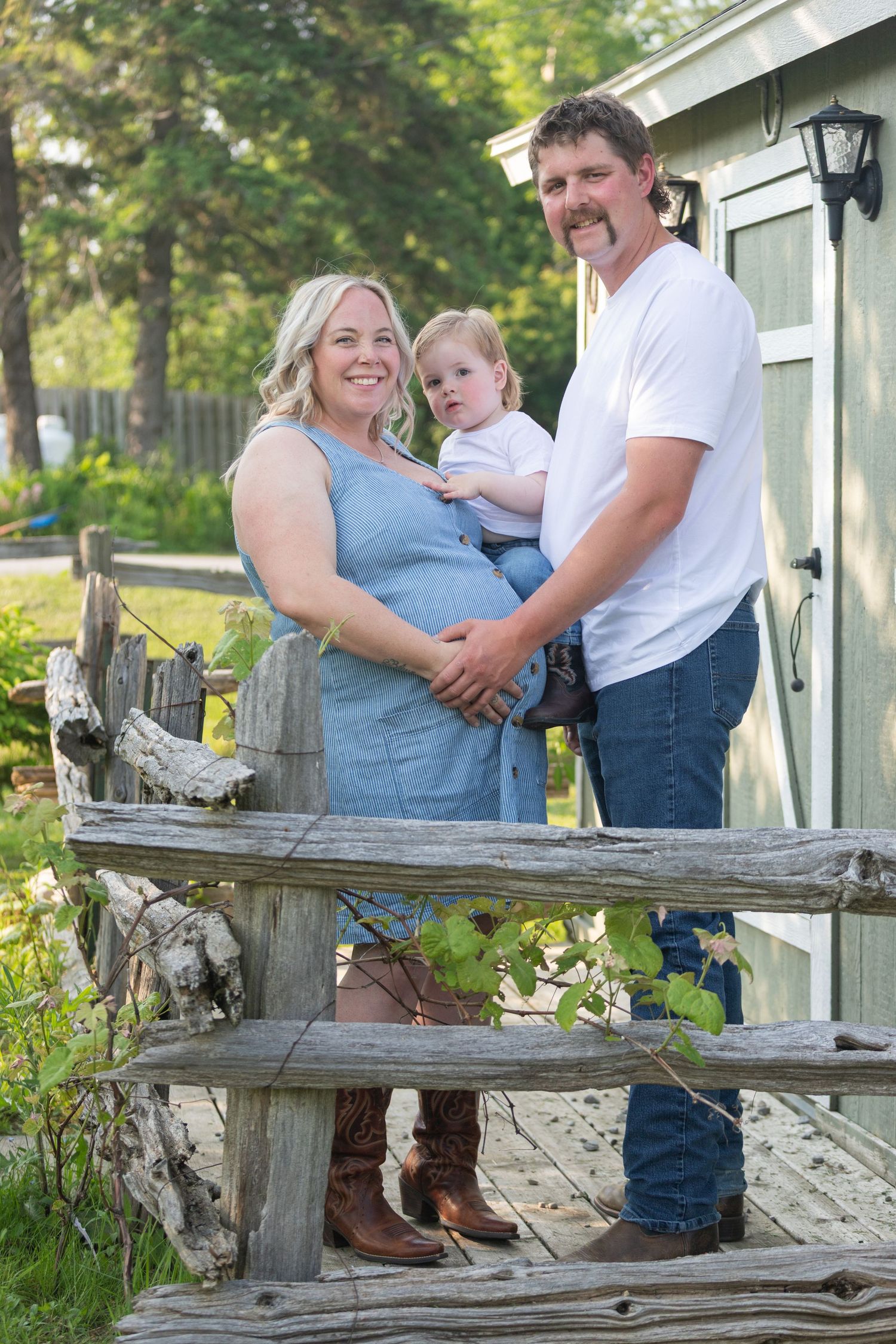 Happy pregnant woman and man holding toddler pose by weathered wooden fence near green shed.